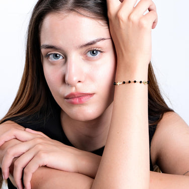 A young woman with light blue eyes and fair skin poses with her head resting on her hand, wearing a 14K gold bracelet with black onyx beads, a cross charm, and a small medallion.