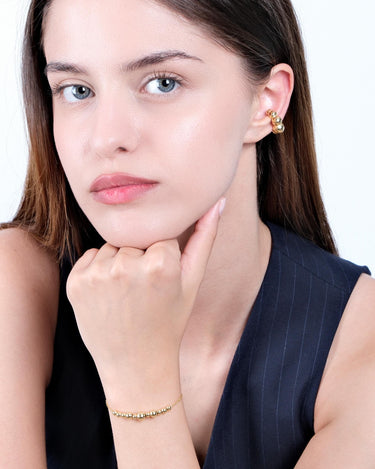 A woman resting her chin on her hand while wearing an 18K Gold Vermeil beaded bracelet, with matching earrings visible.