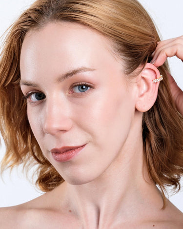 A close-up of a woman with fair skin and light brown hair wearing a gold ear cuff with clear stones. She is gently touching her ear, highlighting the cuff&#39;s placement on the upper ear.