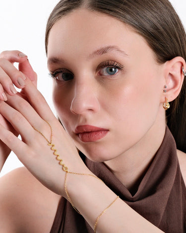 Portrait of a woman with light brown hair, holding her hands near her face, wearing a gold snake hand chain bracelet that features a zigzag pattern.