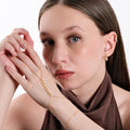 Portrait of a woman with light brown hair, holding her hands near her face, wearing a gold snake hand chain bracelet that features a zigzag pattern.