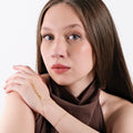 A woman with long brown hair, gazing forward, while her hands are positioned to highlight the gold snake hand chain bracelet with a zigzag design draped over her hand.