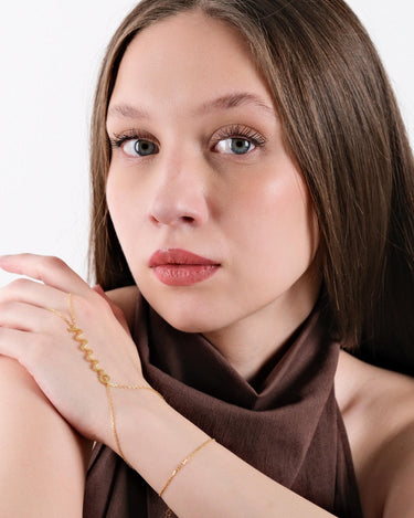 A woman with long brown hair, gazing forward, while her hands are positioned to highlight the gold snake hand chain bracelet with a zigzag design draped over her hand.