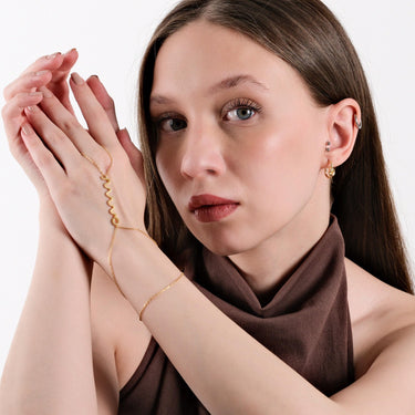 A woman with a serious expression, posing with her hands near her face, displaying the gold snake hand chain bracelet with a zigzag design prominently.