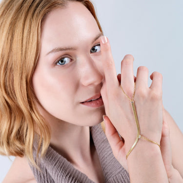 Close-up of a woman with blonde hair, softly smiling while holding her right hand, adorned with a gold vermeil herringbone hand chain bracelet, close to her face.