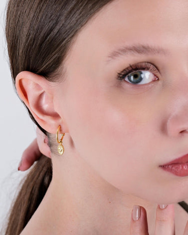 Partial view of a woman&#39;s face wearing a gold vermeil North Star and evil eye earring. The earring is an oval-shaped pendant hanging from a small hoop, and she is holding her hair away from her face.