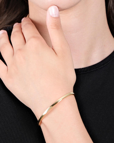 Close-up of a person’s hand gently touching their neck, wearing a thin, shiny gold vermeil bangle bracelet on their wrist. The person is dressed in a black top.