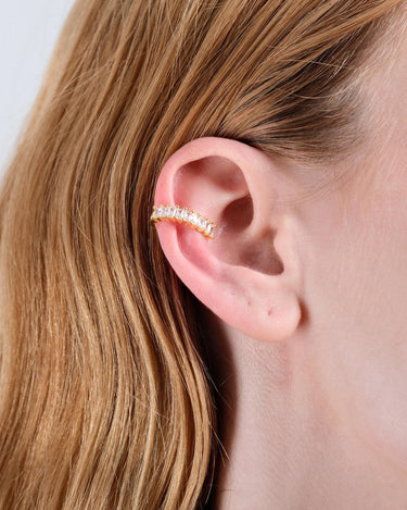 A side view of a woman&#39;s ear adorned with a gold ear cuff featuring clear stones. Her light brown hair is tucked behind the ear, showcasing the cuff&#39;s placement.