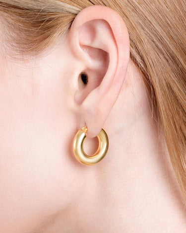 A detailed close-up of a person&#39;s ear showing a shiny, thick 18K gold vermeil chunky hoop earring, highlighting its smooth, rounded shape against light skin and blonde hair.