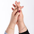 A romantic shot of two hands clasped together, showing a thin gold vermeil bracelet on one wrist and a silver bracelet on the other, each with a delicate loop centerpiece.