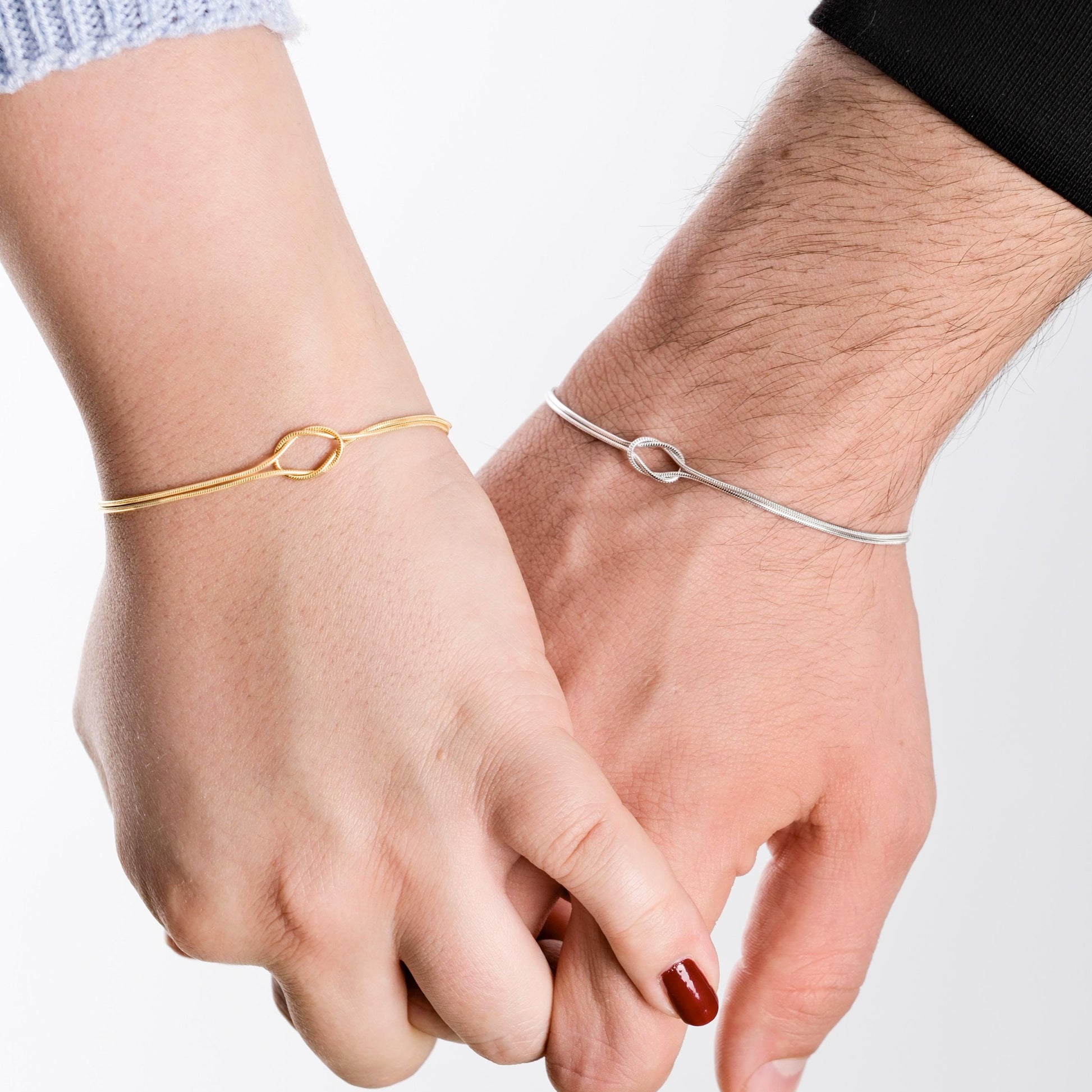 A close-up of two hands gently holding, highlighting a pair of matching bracelets: one in gold vermeil and the other in silver, both designed with a central knot feature.