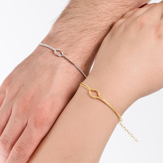 A close-up view of two hands side by side, one wearing a thin gold vermeil bracelet and the other a silver one, both featuring an elegant, minimalist loop design.