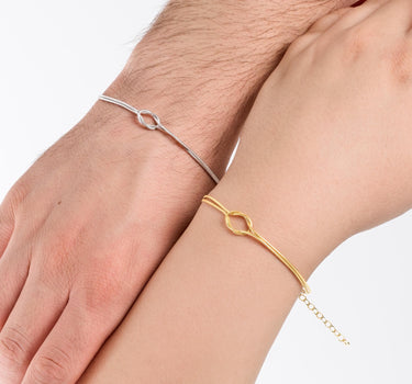 A close-up view of two hands side by side, one wearing a thin gold vermeil bracelet and the other a silver one, both featuring an elegant, minimalist loop design.