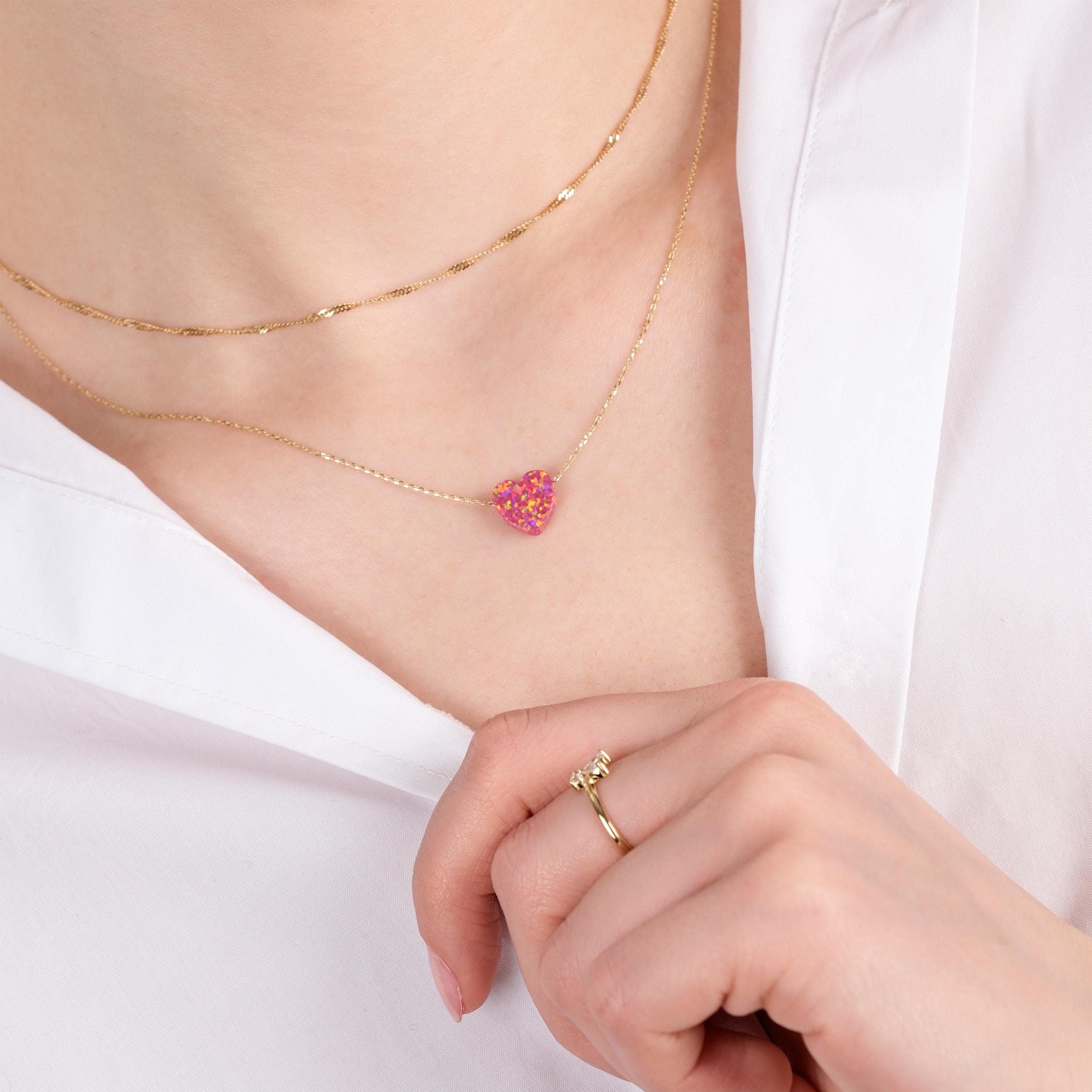 Close-up of a woman wearing a layered 14K solid gold dainty opal heart necklace, gently holding her white shirt collar with a hand adorned by a delicate gold ring.
