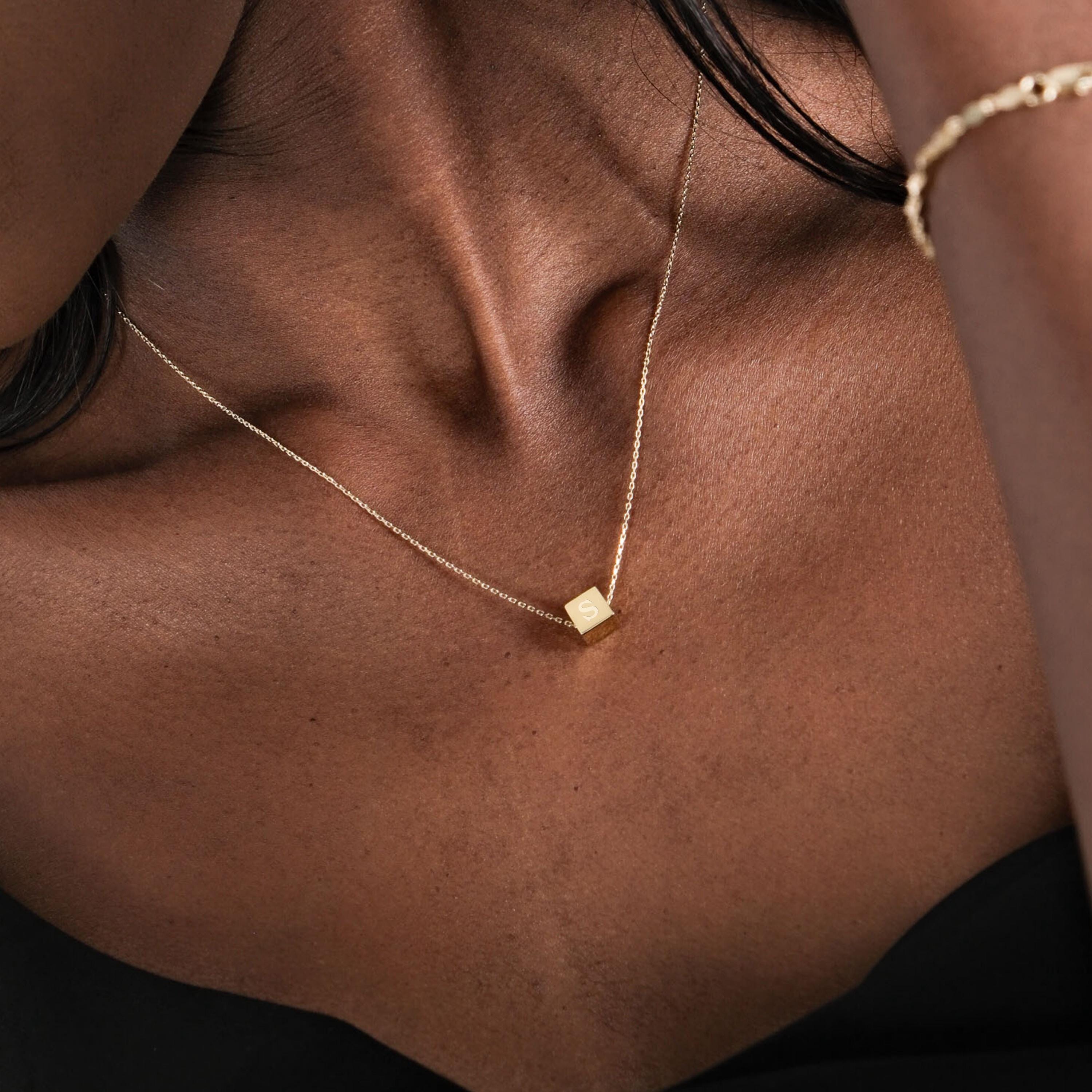 Side angle of a gold cube initial necklace resting on the wearer's collarbone.