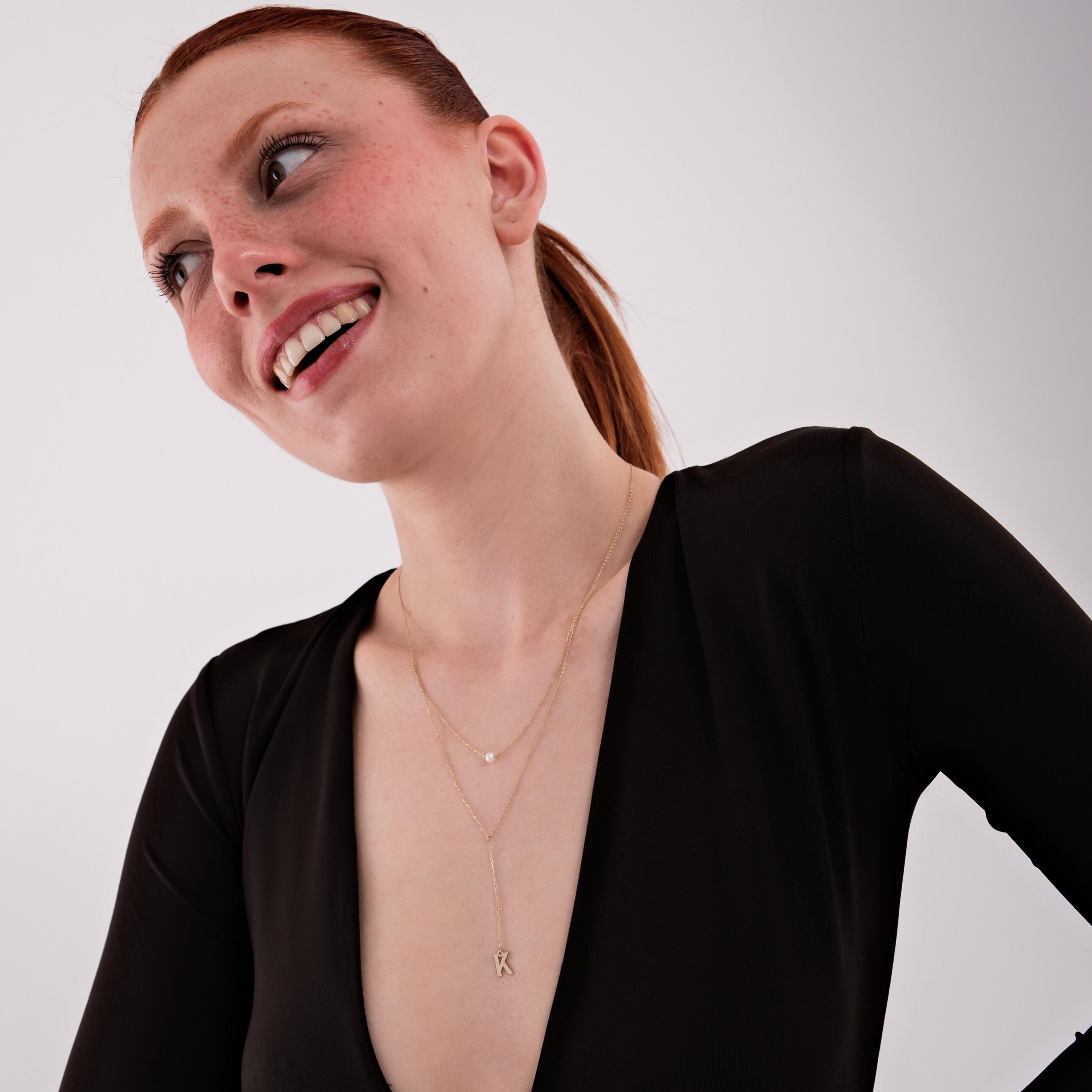 A smiling model wearing a dainty 14K solid gold pearl lariat necklace with layered chains, one with a pearl and the other with a letter 'K' drop.
