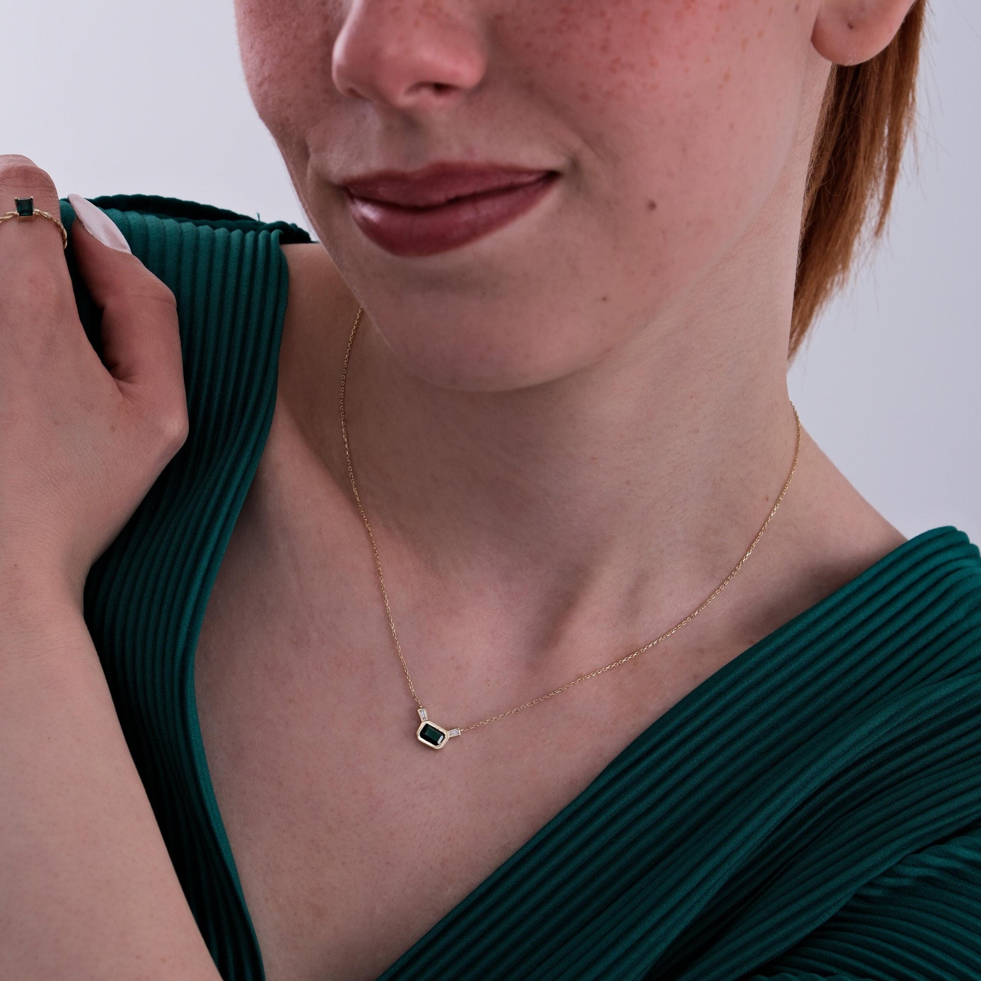 A woman gently touches her shoulder while wearing the 14K Solid Gold Emerald Cut Necklace, the deep green stone catching light subtly.