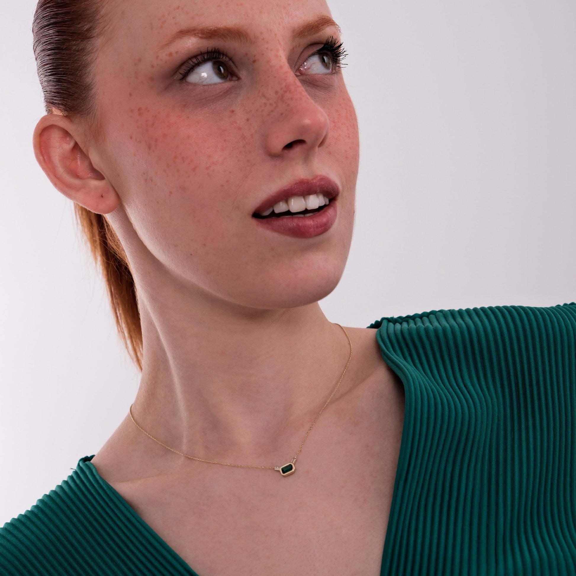 Looking upward, the model wears a 14K Solid Gold Emerald Cut Necklace that glimmers softly against her skin and emerald-toned blouse.
