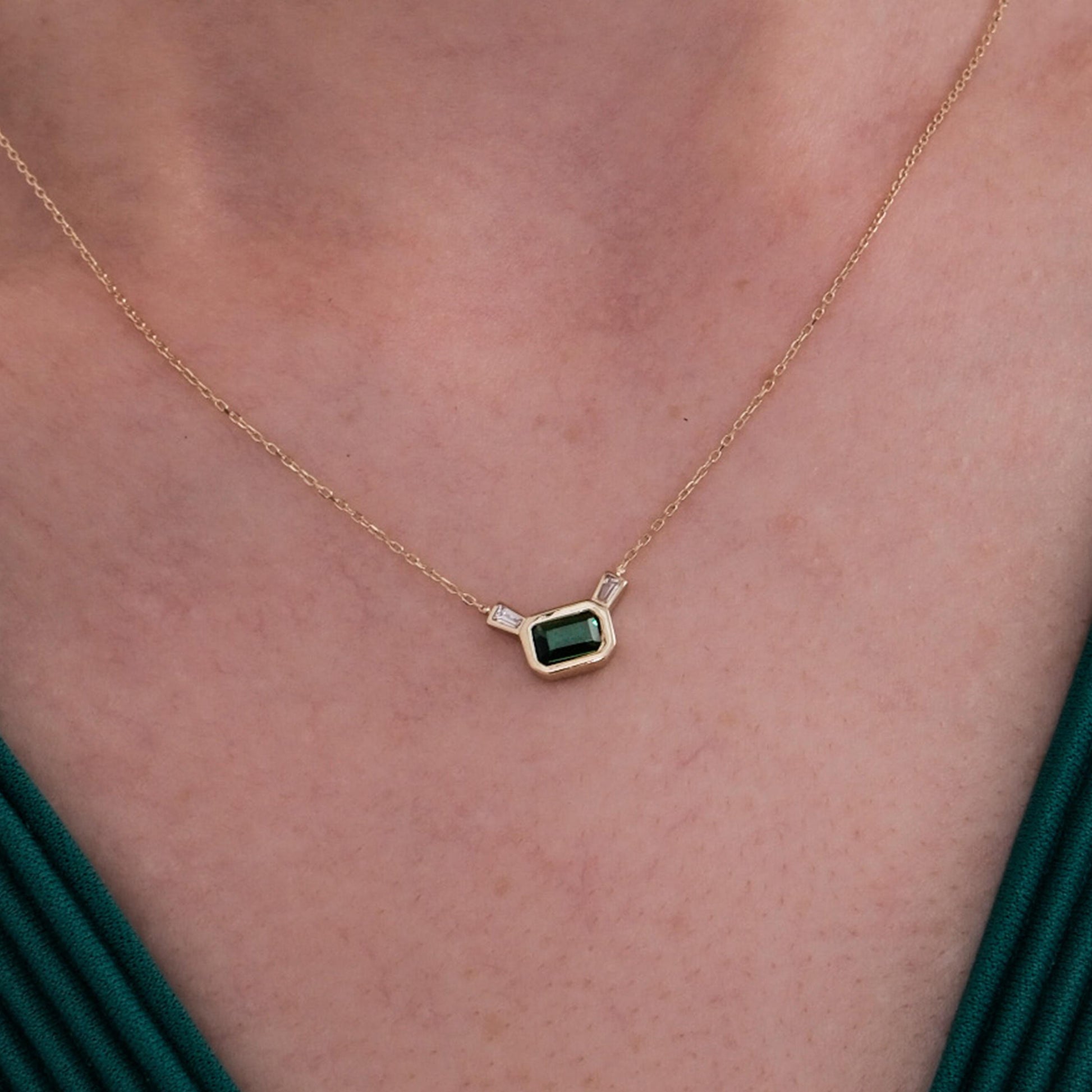 Close-up shot of a 14K Solid Gold Emerald Cut Necklace resting at the center of a woman&#39;s collarbone, showcasing its rich green gemstone and fine gold chain.