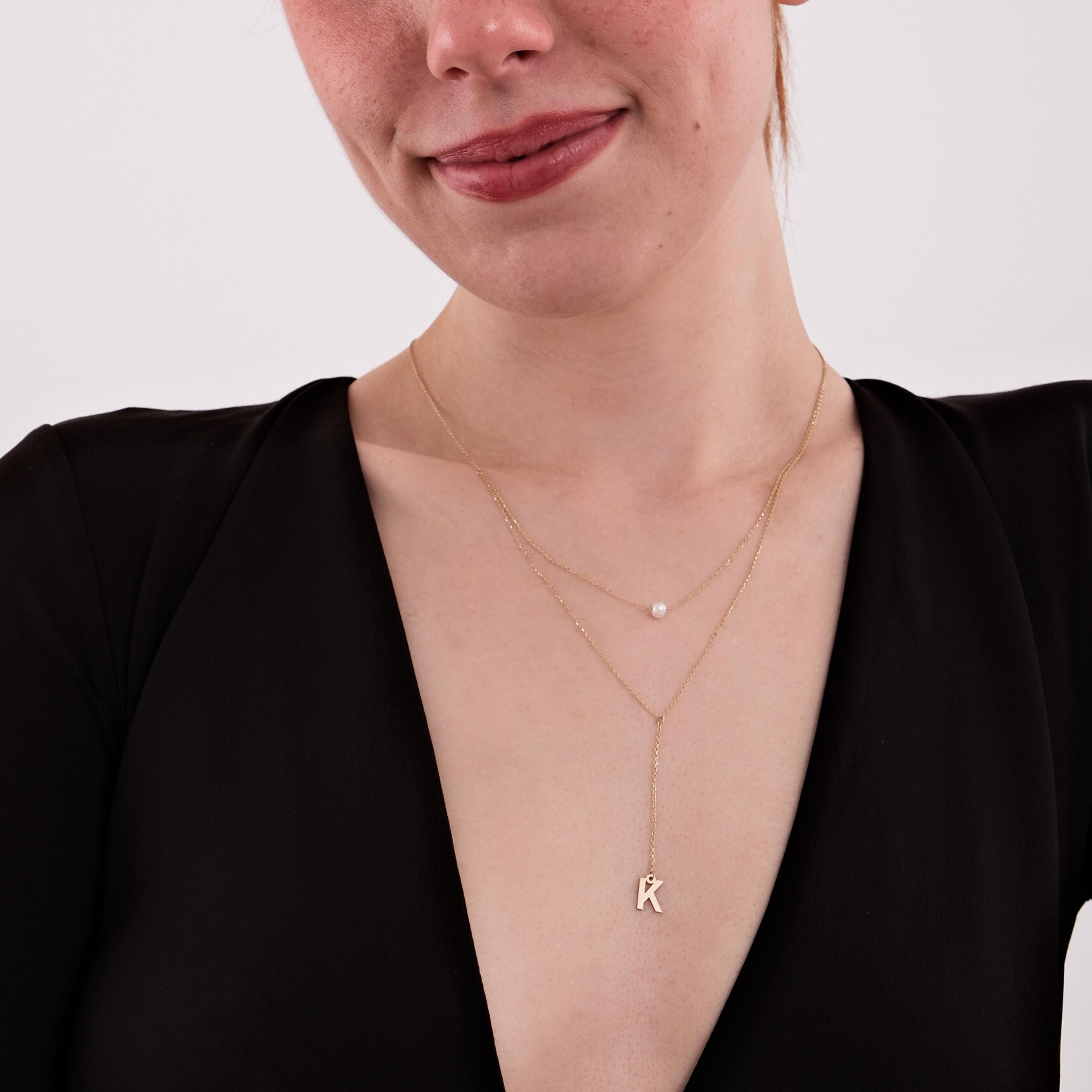 Close-up of a woman wearing a 14K solid gold pearl lariat necklace with a pearl detail and a hanging letter 'K' pendant, styled with a deep V-neck black blouse.