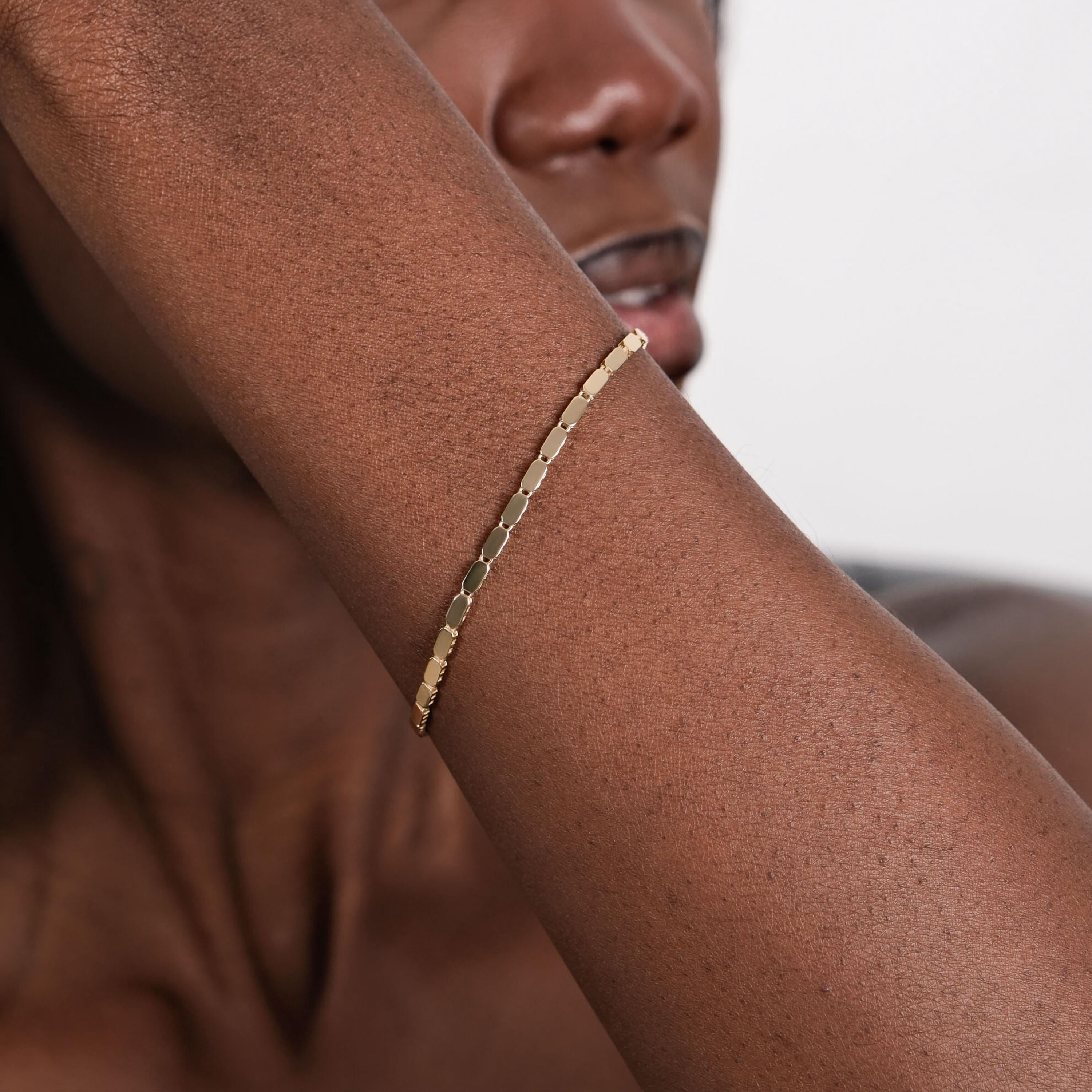 A close-up of a wrist wearing a sleek 14K gold mirror chain bracelet with reflective, rectangular links.
