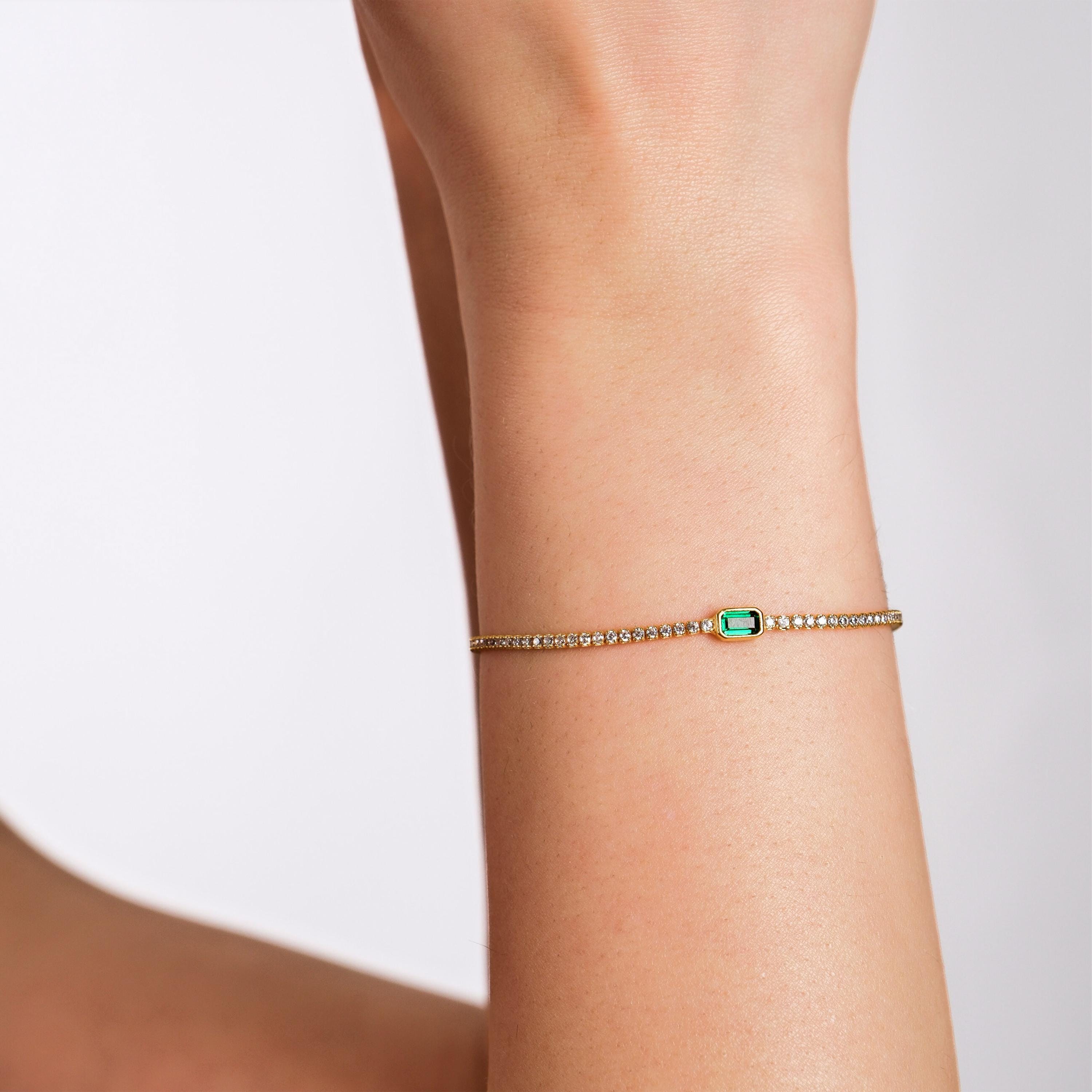 A close-up of a wrist wearing an 18K gold vermeil emerald bracelet, featuring a delicate emerald stone centerpiece.