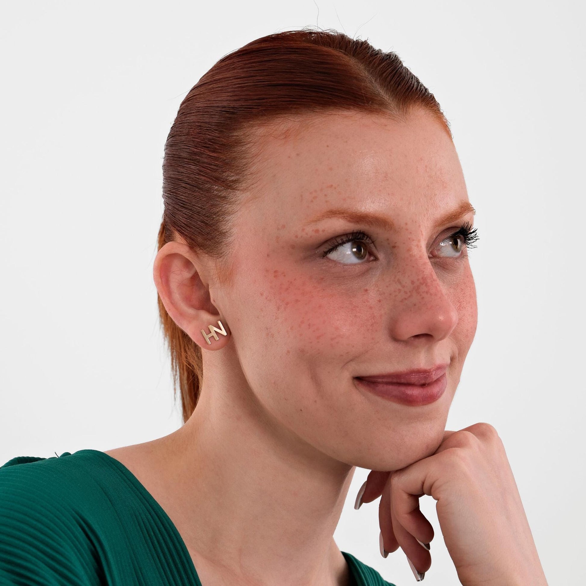 Smiling woman resting her chin on her hand while wearing 14K solid gold &quot;HN&quot; letter stud earrings.