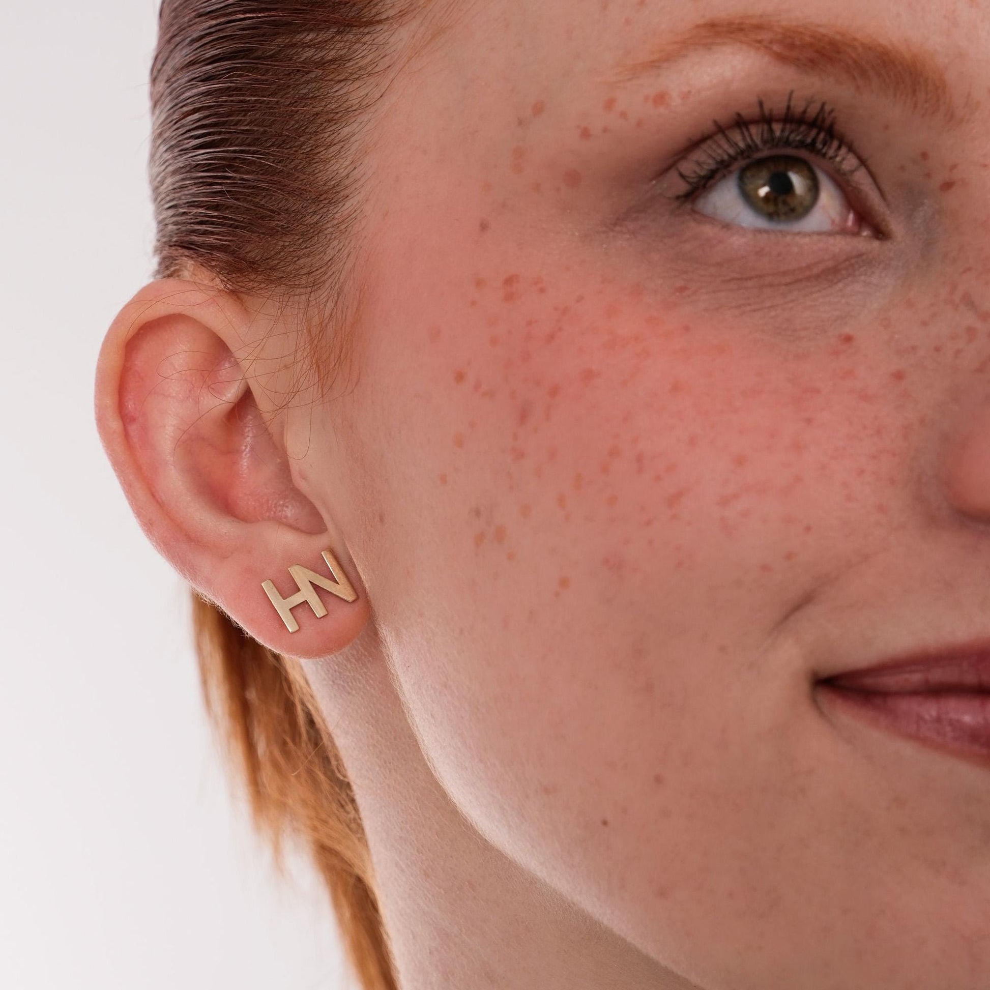 Close-up of a woman’s ear and cheek, showing 14K solid gold initial earrings with the letters &quot;HN&quot;.