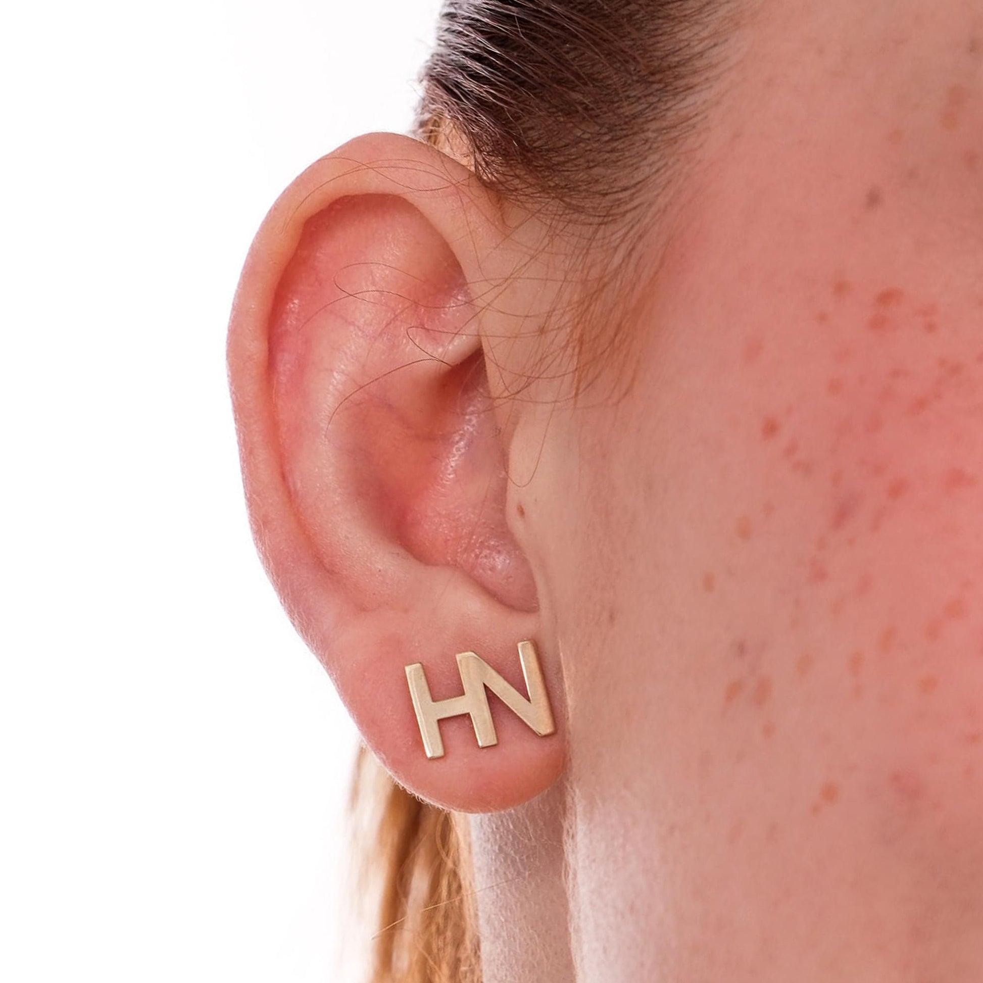 Close-up of an ear wearing minimalist 14K solid gold letter stud earrings forming the initials &quot;HN&quot;.
