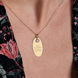A close-up of a woman's neck wearing a 14K solid gold engraved necklace with an oval pendant, featuring the inscription "First My Mother, Forever My Friend" and a small birthstone charm.