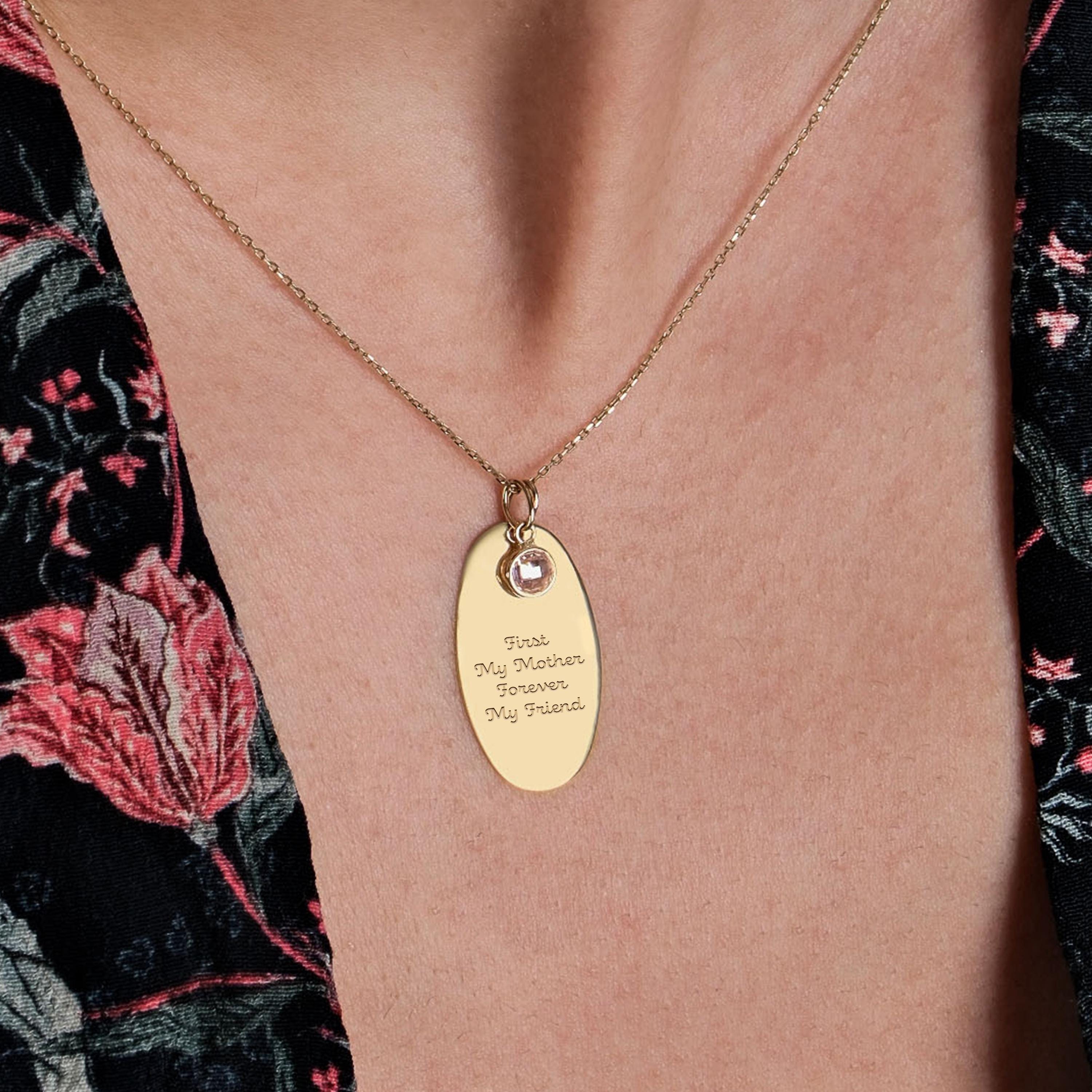 A close-up of a woman's neck wearing a 14K solid gold engraved necklace with an oval pendant, featuring the inscription "First My Mother, Forever My Friend" and a small birthstone charm.