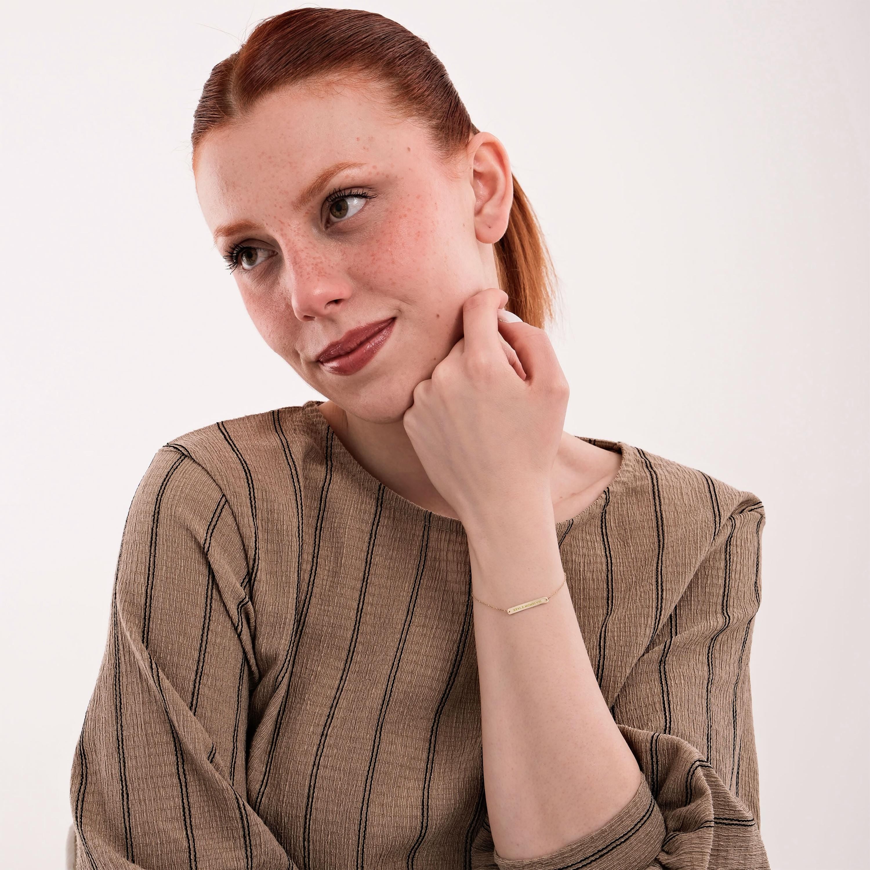 Portrait of a red-haired woman in a striped blouse, subtly displaying a 14K solid gold engraved bar bracelet.