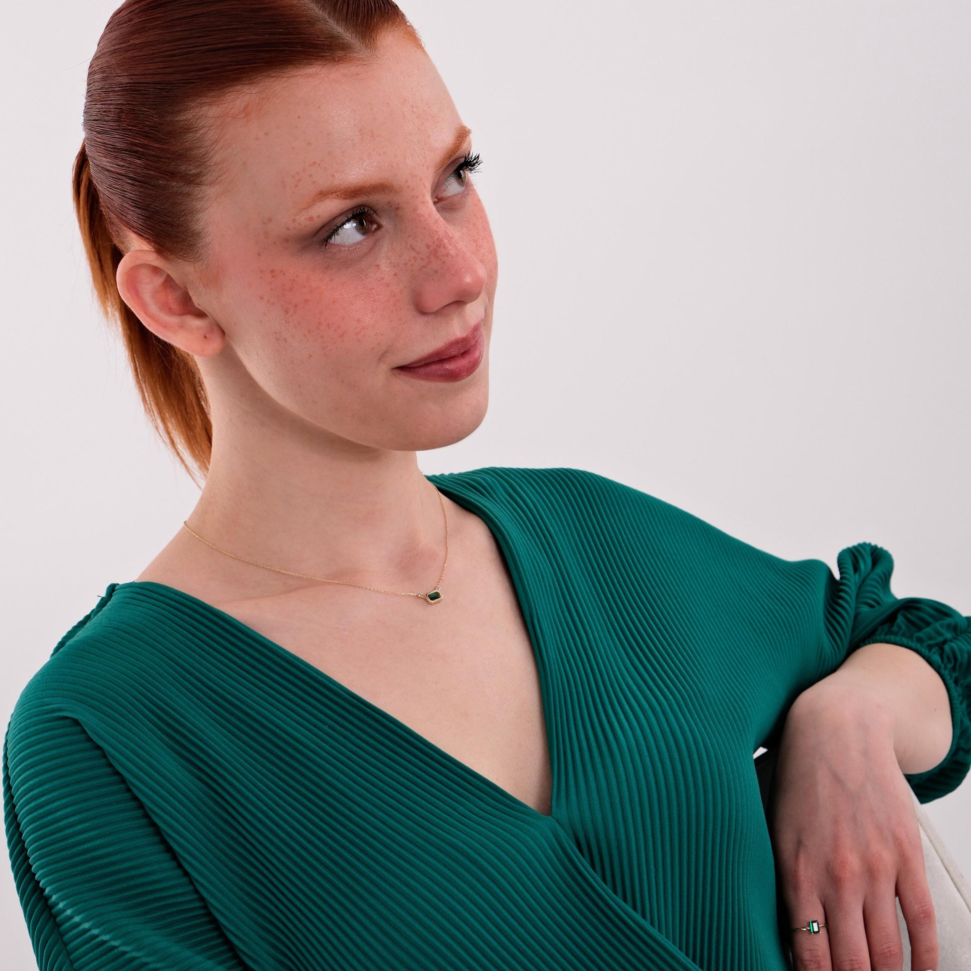The model poses seated, dressed in green pleats and wearing a 14K Solid Gold Emerald Cut Necklace that adds a refined touch to her neckline.