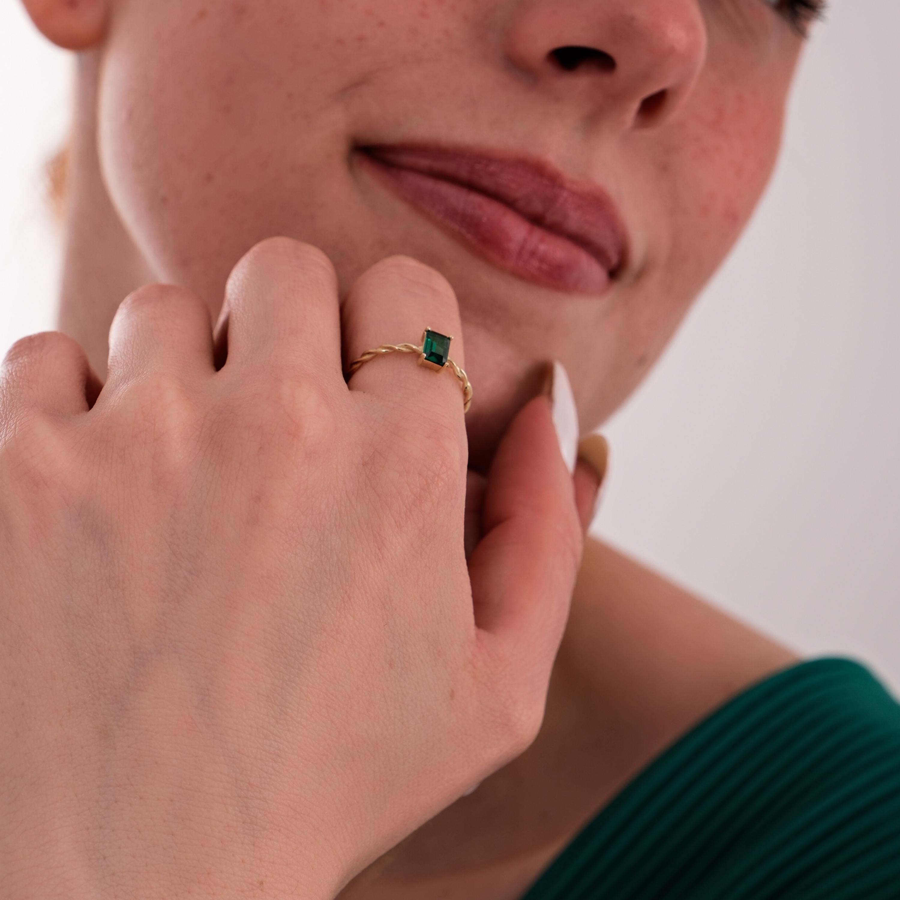 The model rests her hand near her chin, showcasing the 14K Solid Gold Emerald Cut Ring with a twisted band and deep green stone.