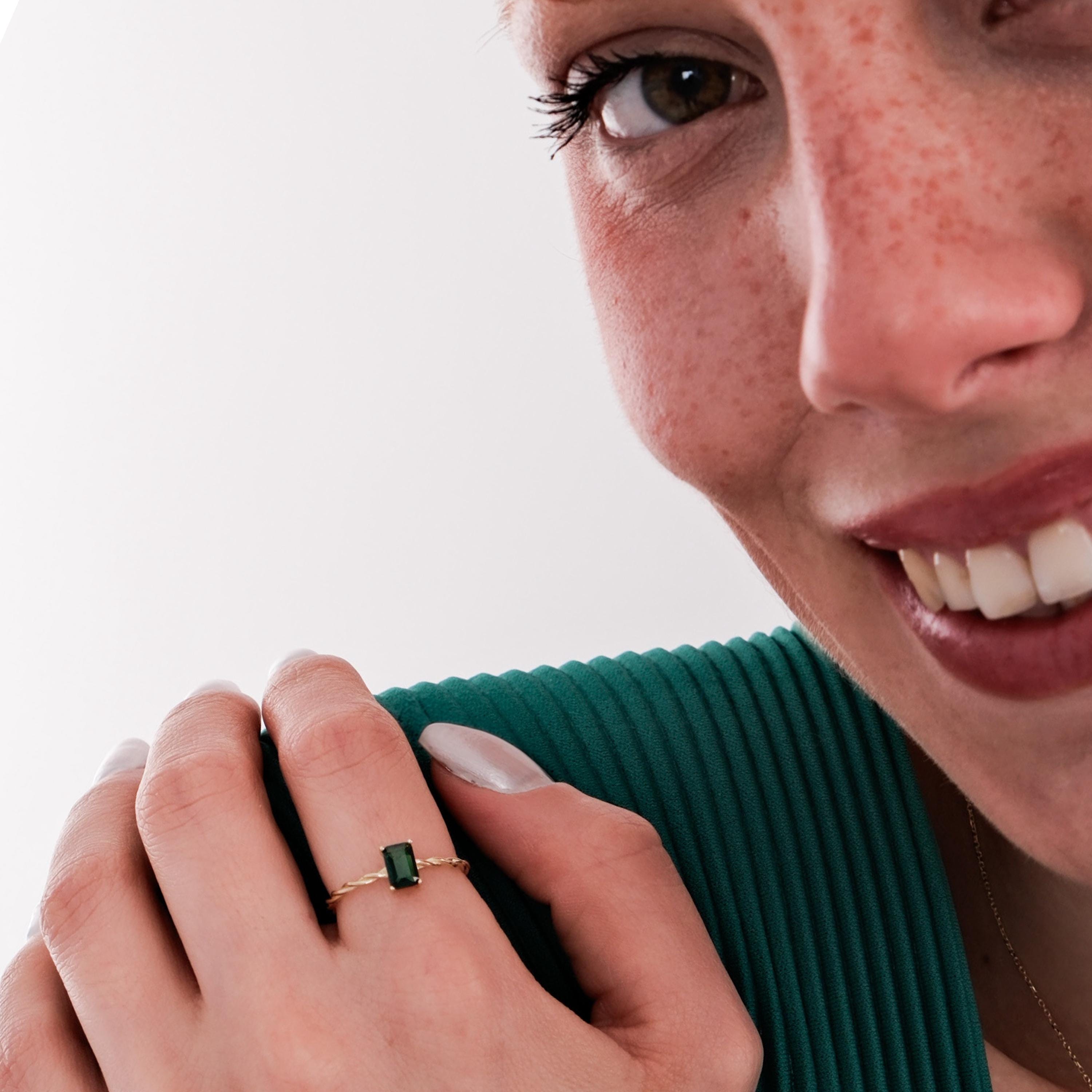 The model smiles while showing off a 14K Solid Gold Emerald Cut Ring with a deep green stone, worn on her index finger.