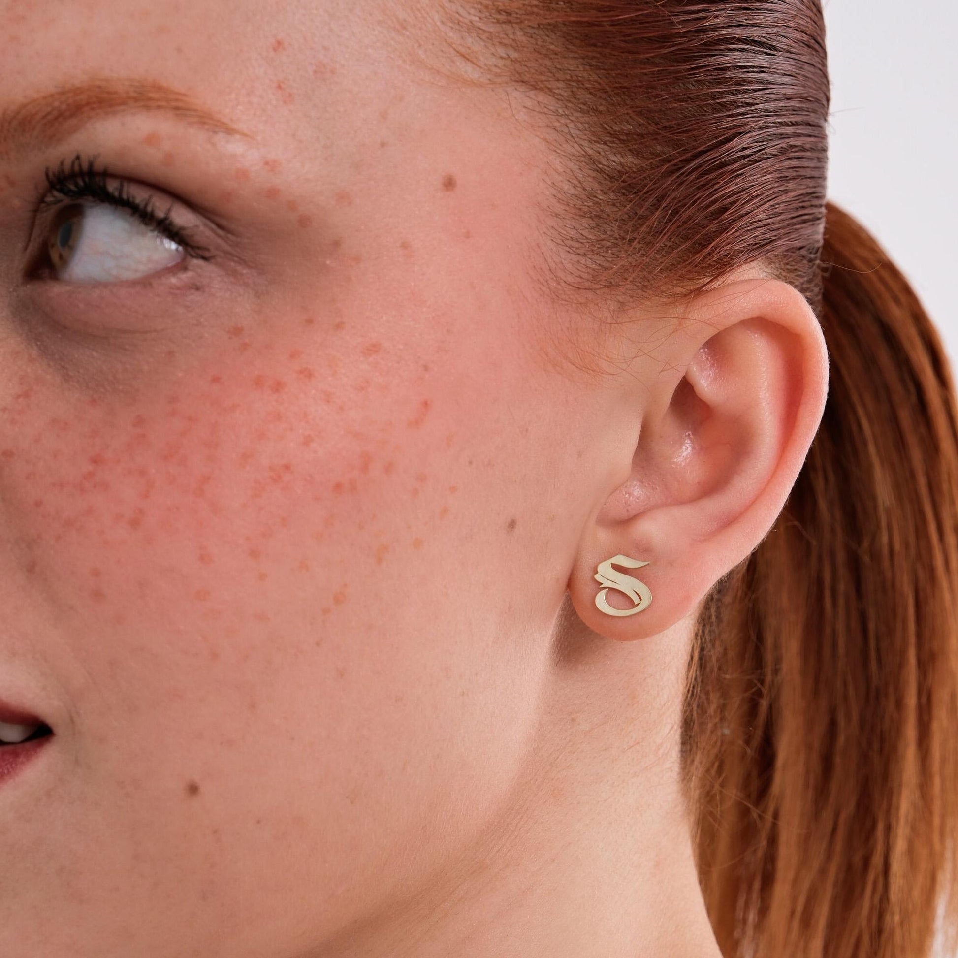 The model gazes to the side, wearing a single 14K Solid Gold Initial Stud Earring shaped like the letter “S”.