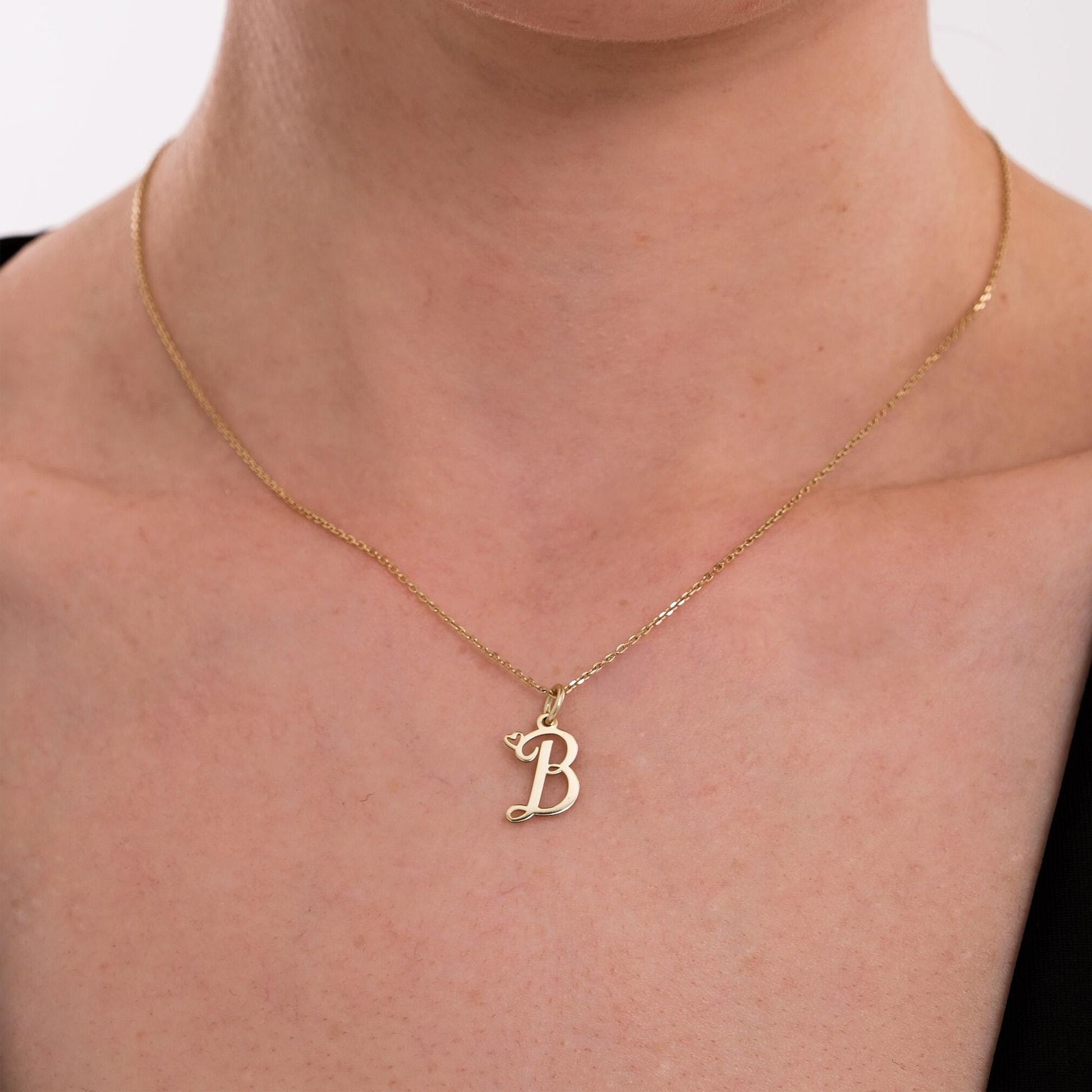 A close-up of a 14K solid gold cursive initial necklace featuring the letter &quot;B&quot; and a tiny heart charm.