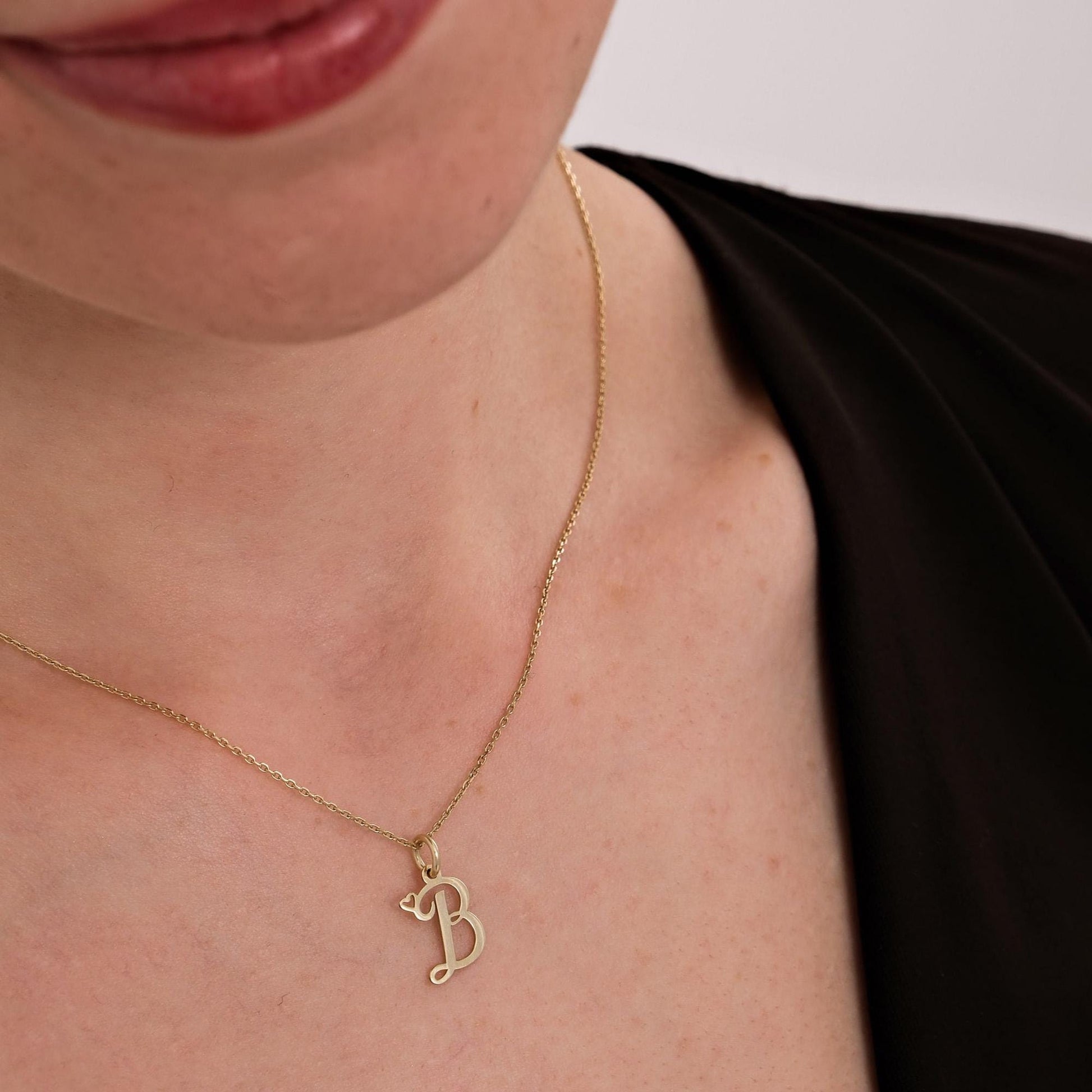 A woman wears a 14K solid gold cursive initial necklace with the &quot;B&quot; pendant styled over a black V-neck top.