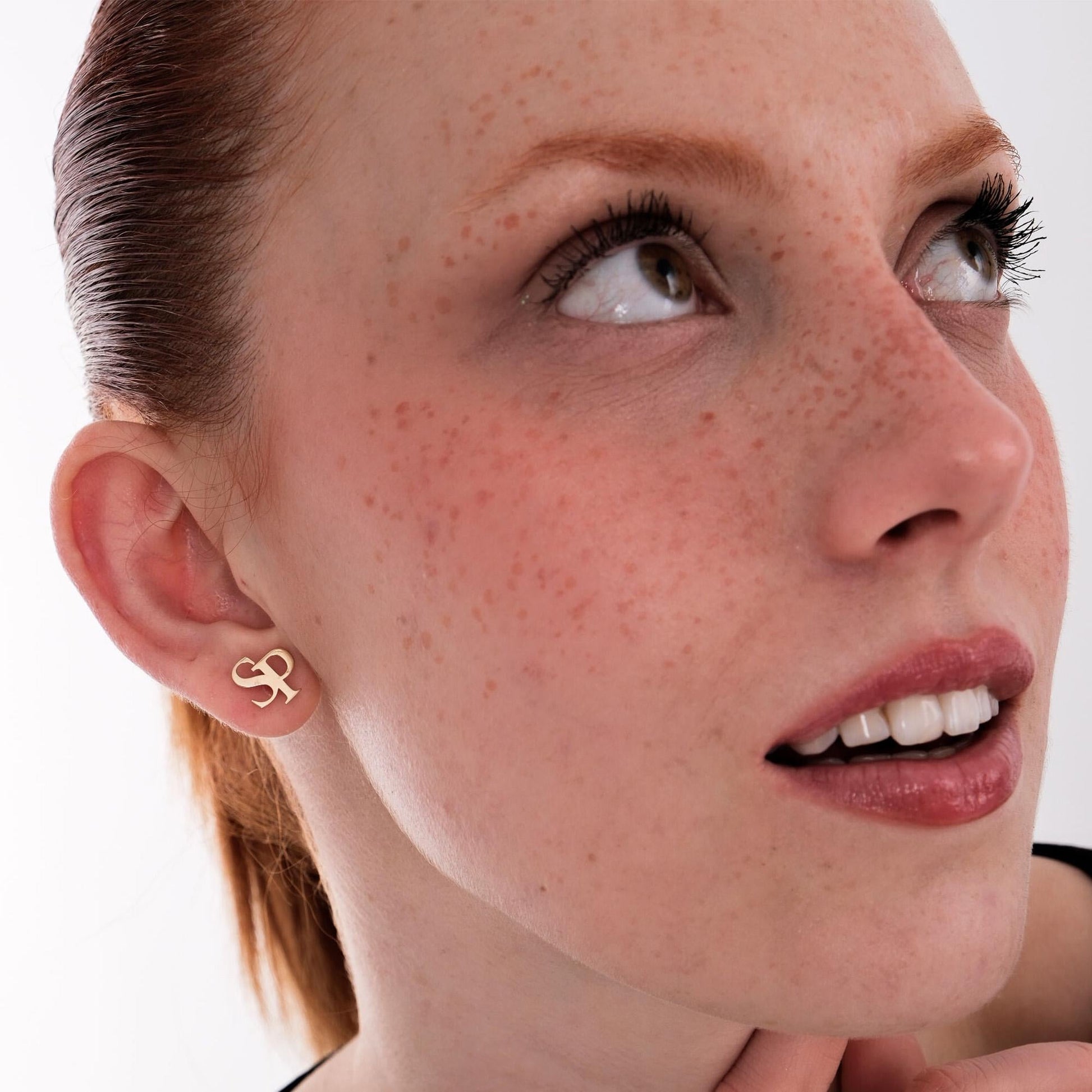 Woman with freckles wearing 14K solid gold intertwined initial earrings shaped as “S” and “P”, looking upwards with a soft smile.