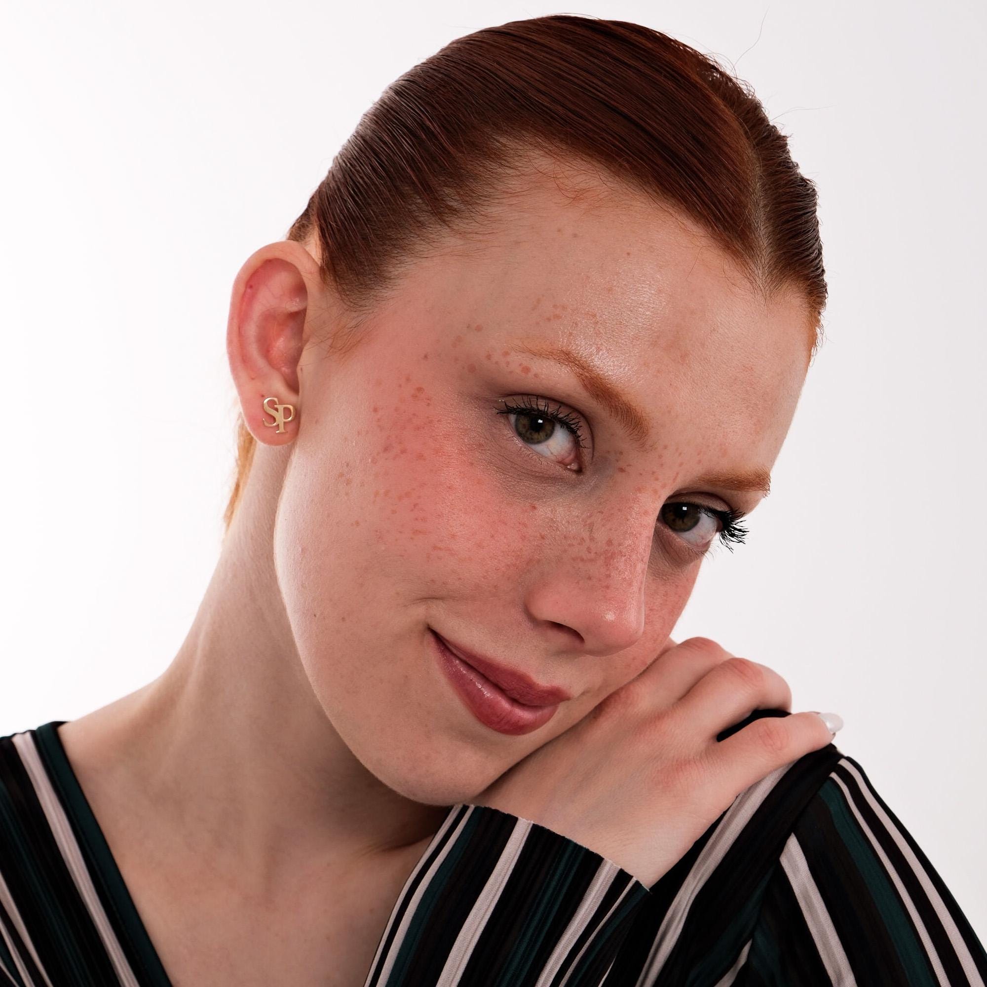 Portrait of a smiling woman with tied-back hair wearing 14K solid gold intertwined initial earrings, resting her head gently on her hands.