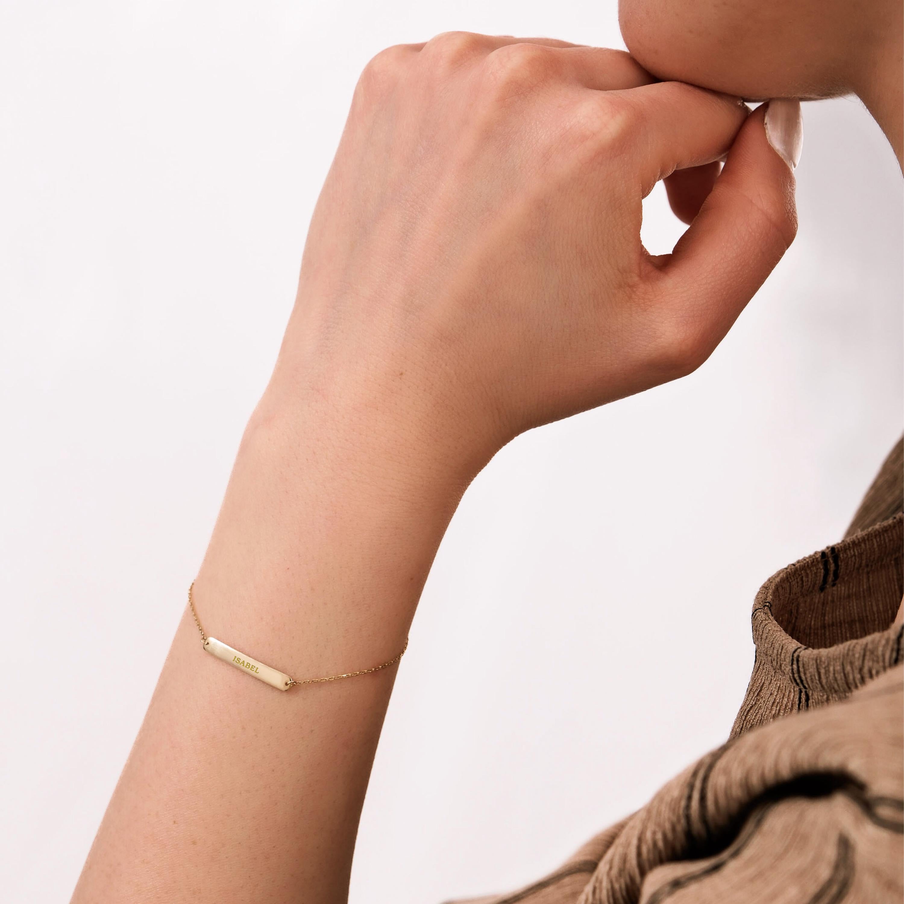 Profile view of a woman's wrist featuring a delicate 14K solid gold custom name bracelet engraved with "ISABEL" against a light background.