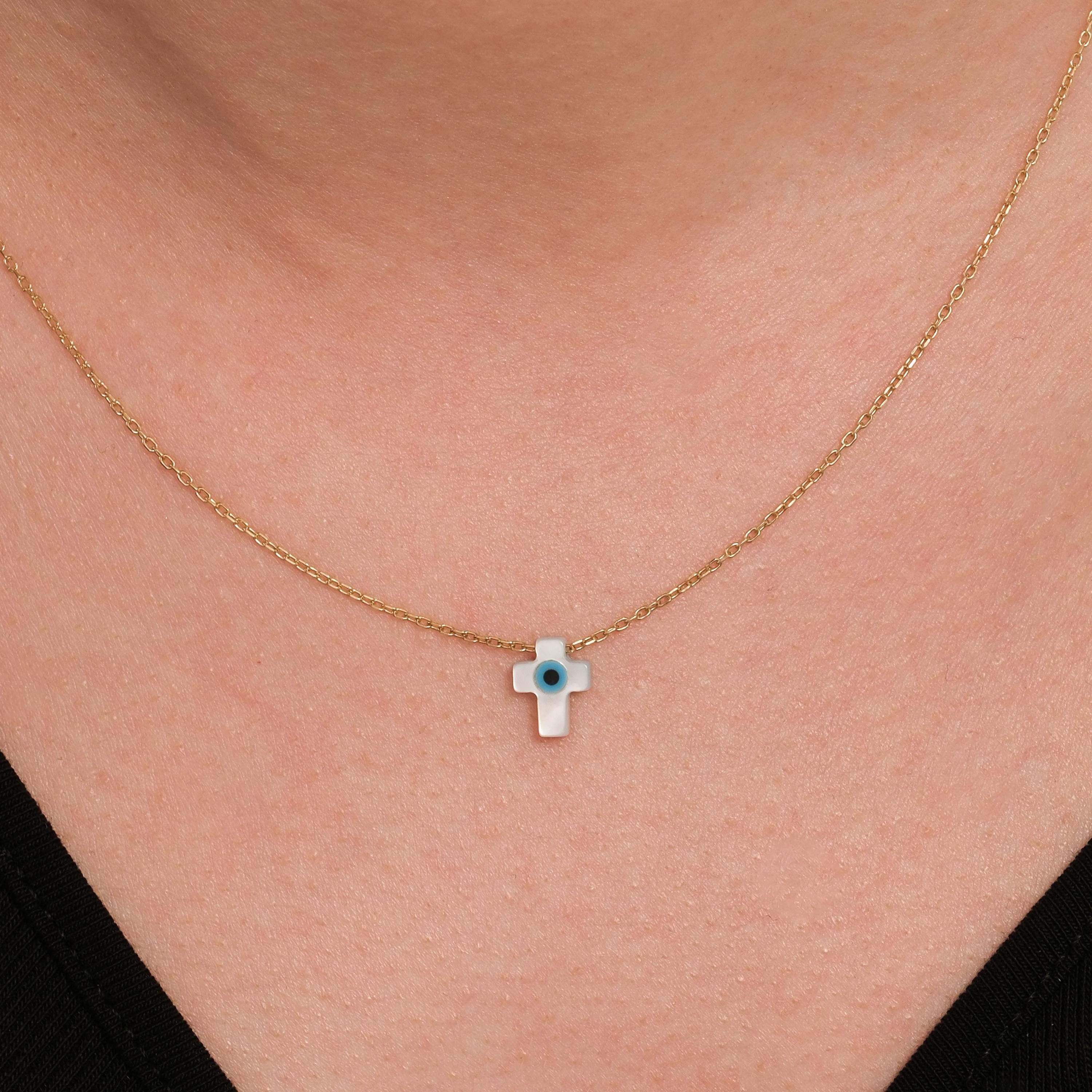Close-up of a woman wearing a 14K solid gold cross evil eye necklace with a glossy white charm and a blue center.