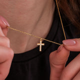 Woman gently holding a white-stone cross pendant necklace in 18K gold vermeil close to her neck.