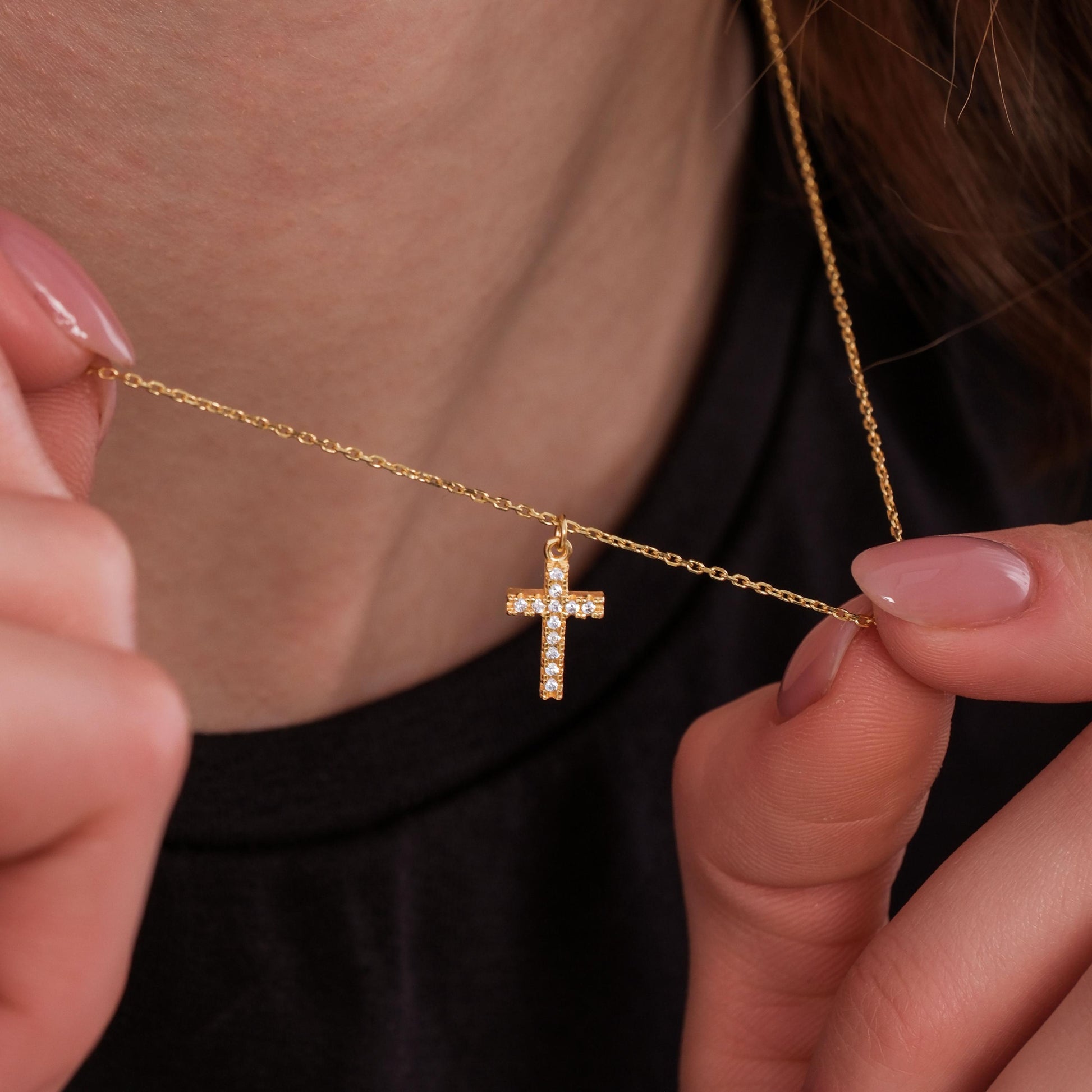 Woman gently holding a white-stone cross pendant necklace in 18K gold vermeil close to her neck.