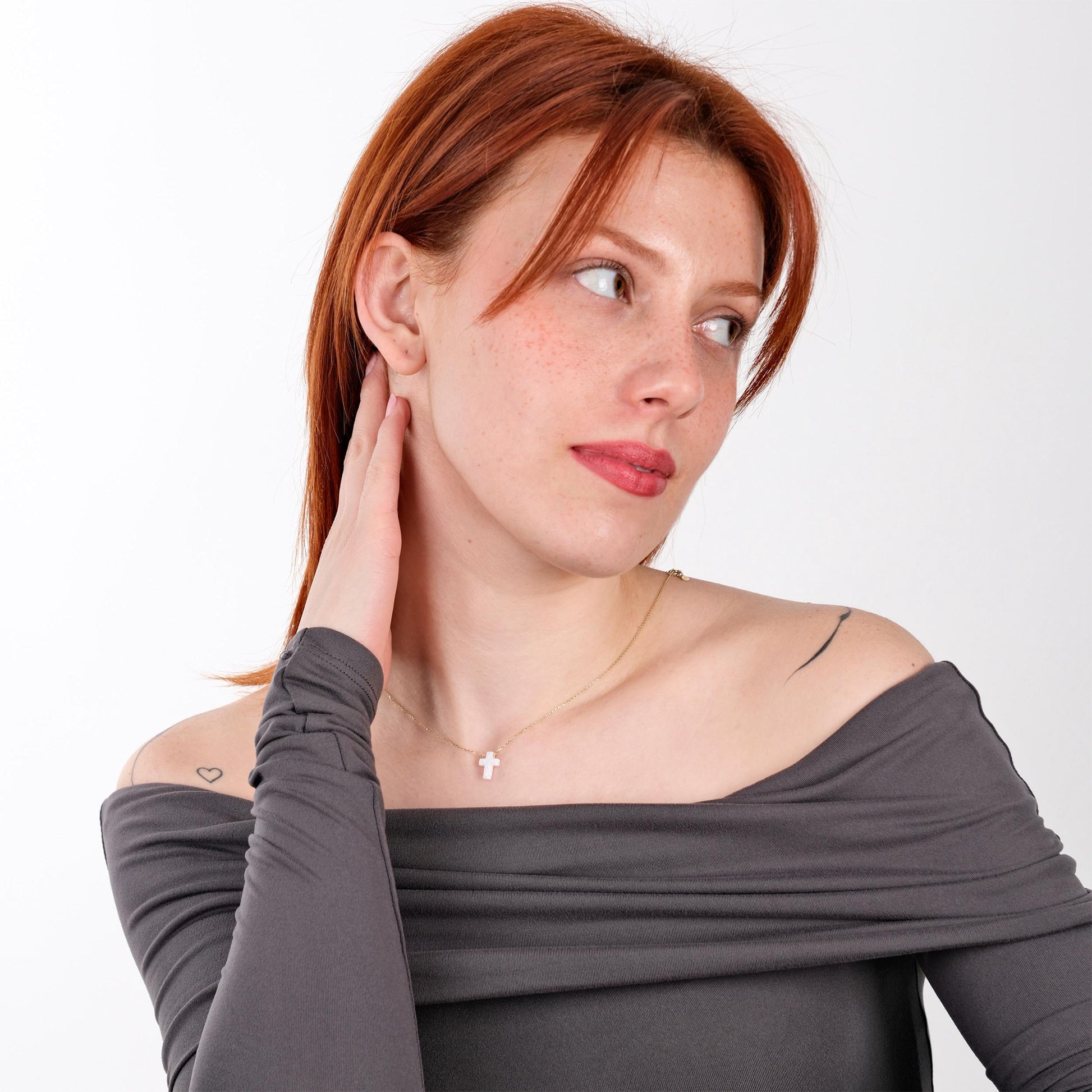 Woman posing in an off-shoulder gray top while wearing a 14K solid gold white opal cross necklace.