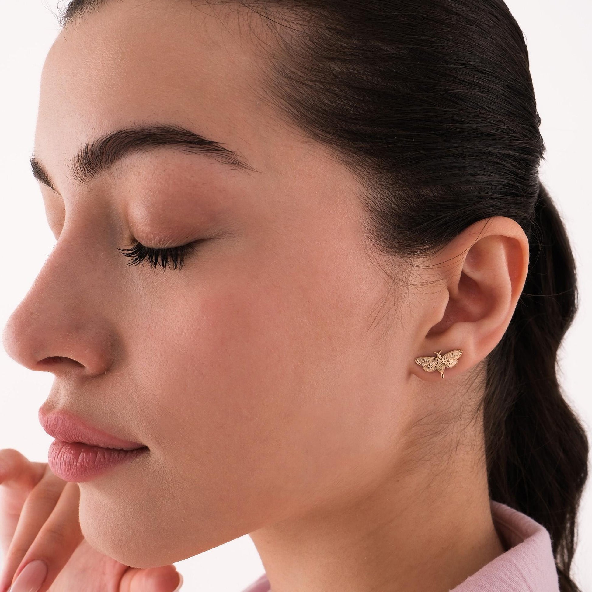 Side view of a woman with closed eyes wearing 14K solid gold moth earrings, styled with a soft pink top.