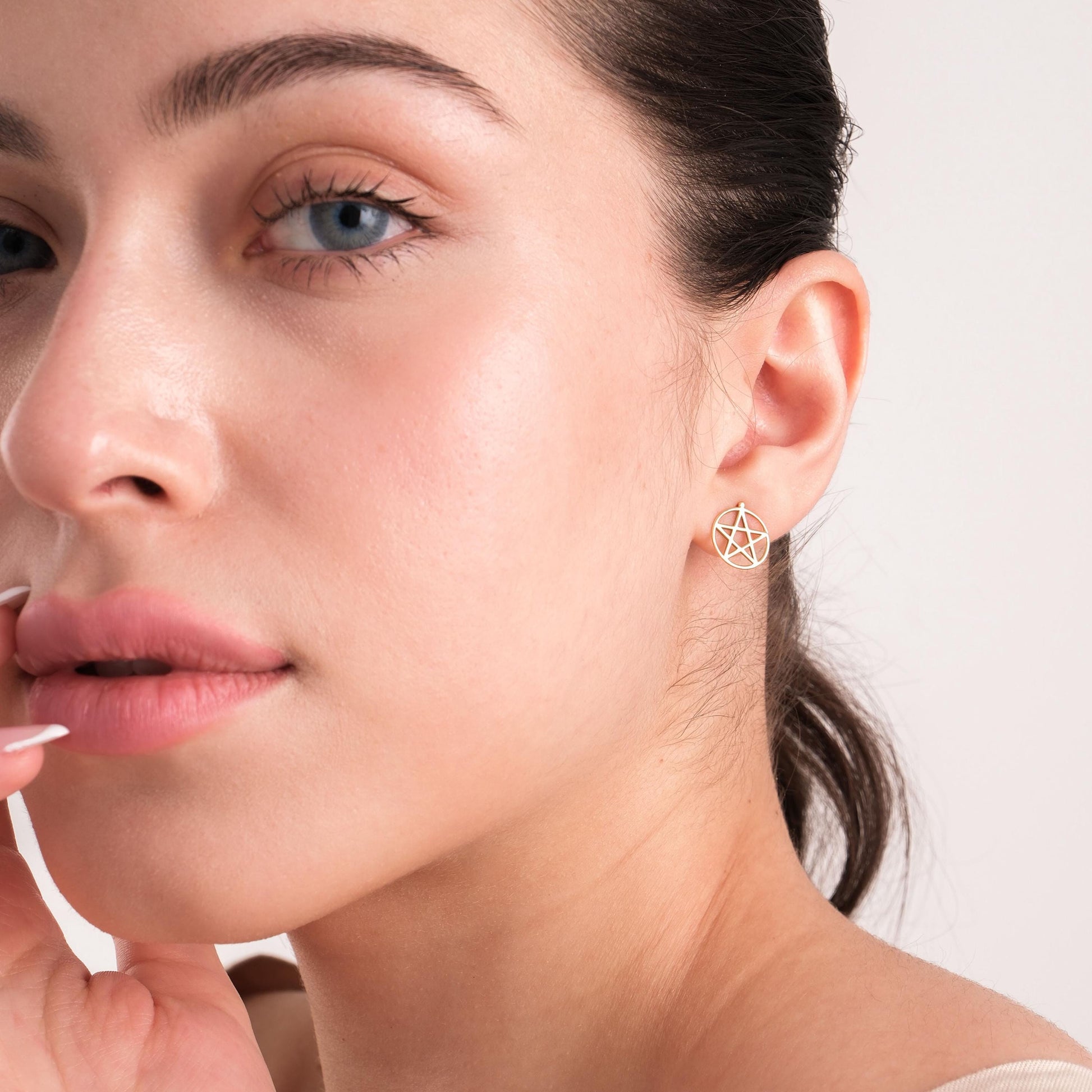 Portrait of a woman gently holding her face while wearing a 14K solid gold pentagram stud earring.