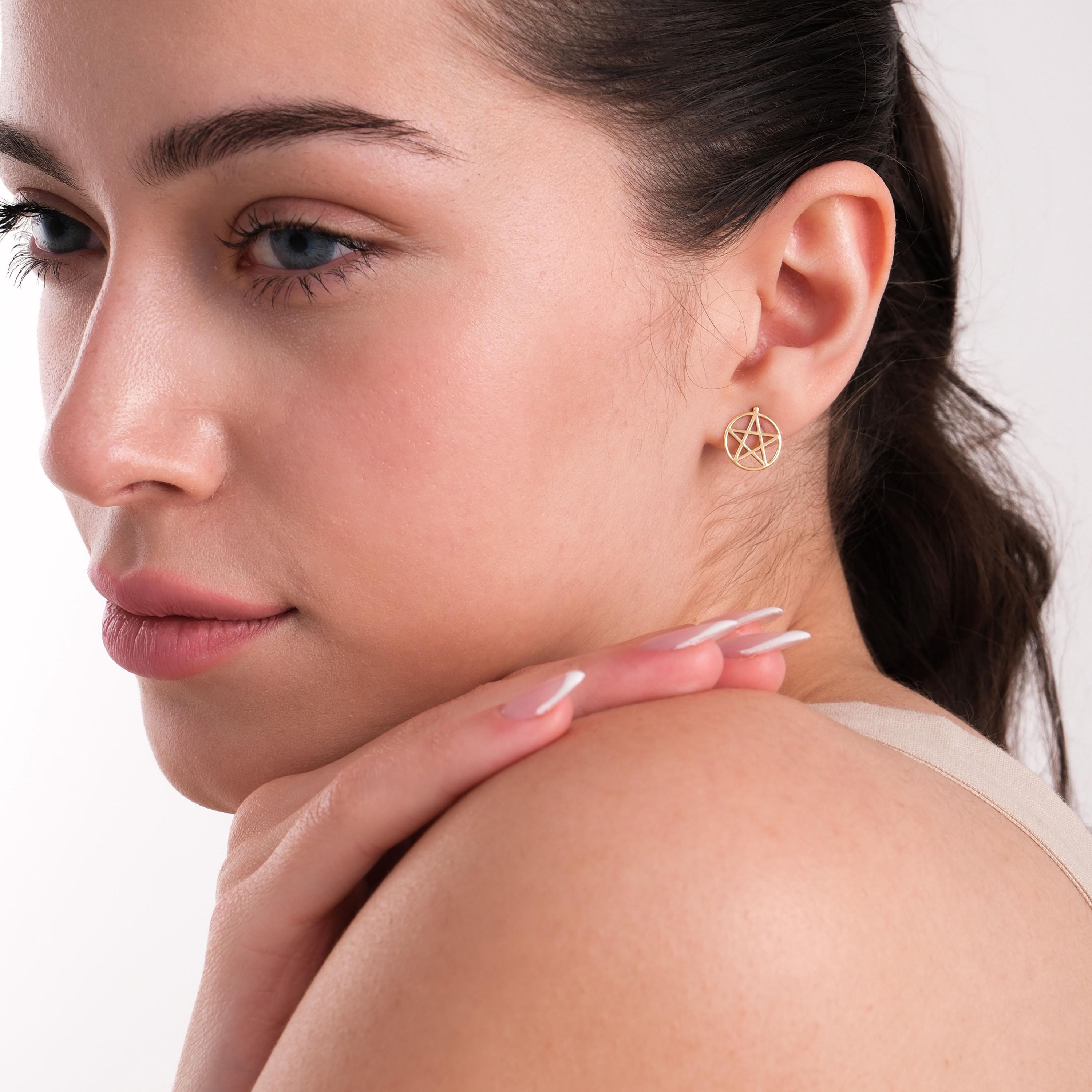 A woman in profile, resting her hand on her shoulder while displaying a 14K solid gold pentagram stud earring.