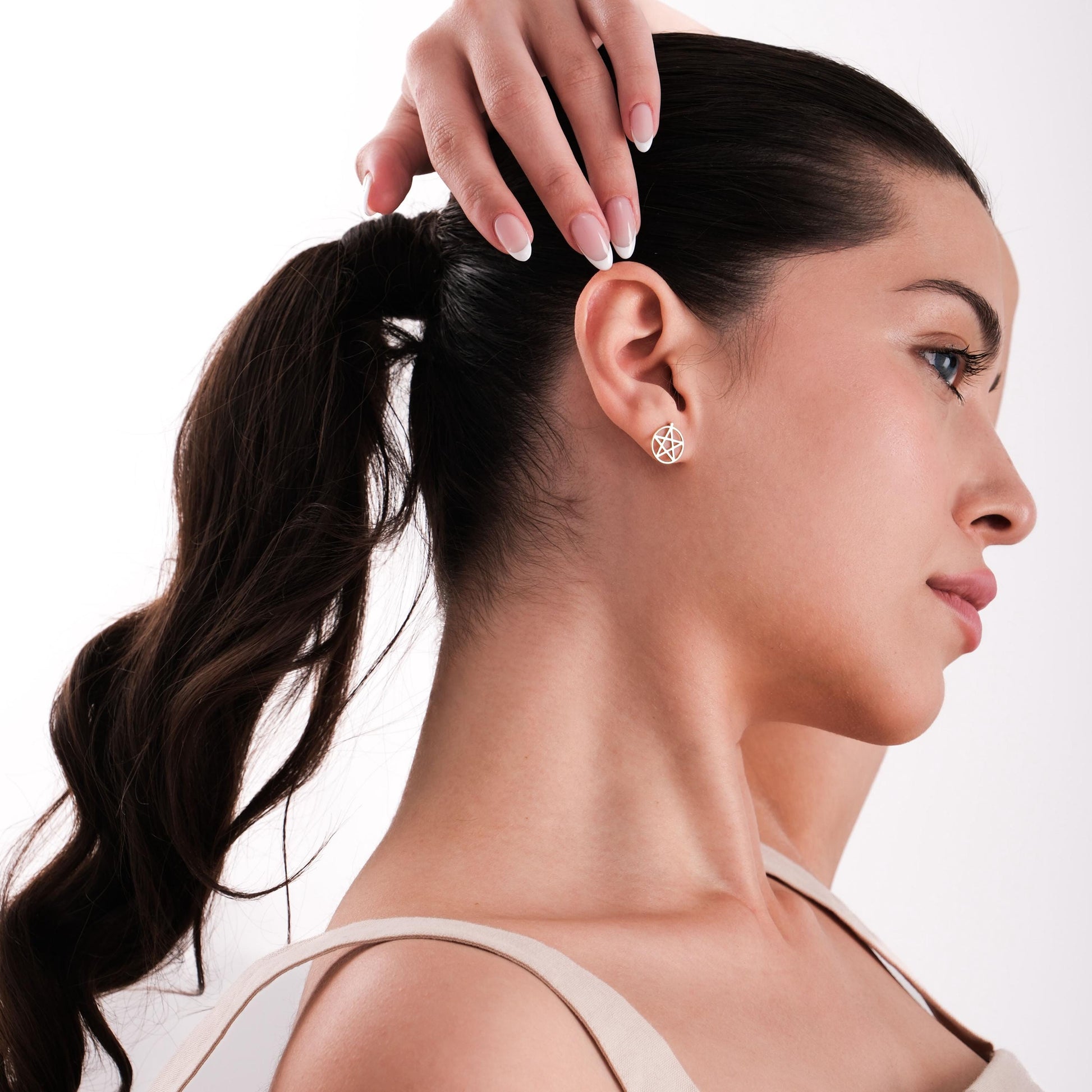 Side view of a woman with a ponytail wearing a 14K solid gold pentagram stud earring, touching her hair.