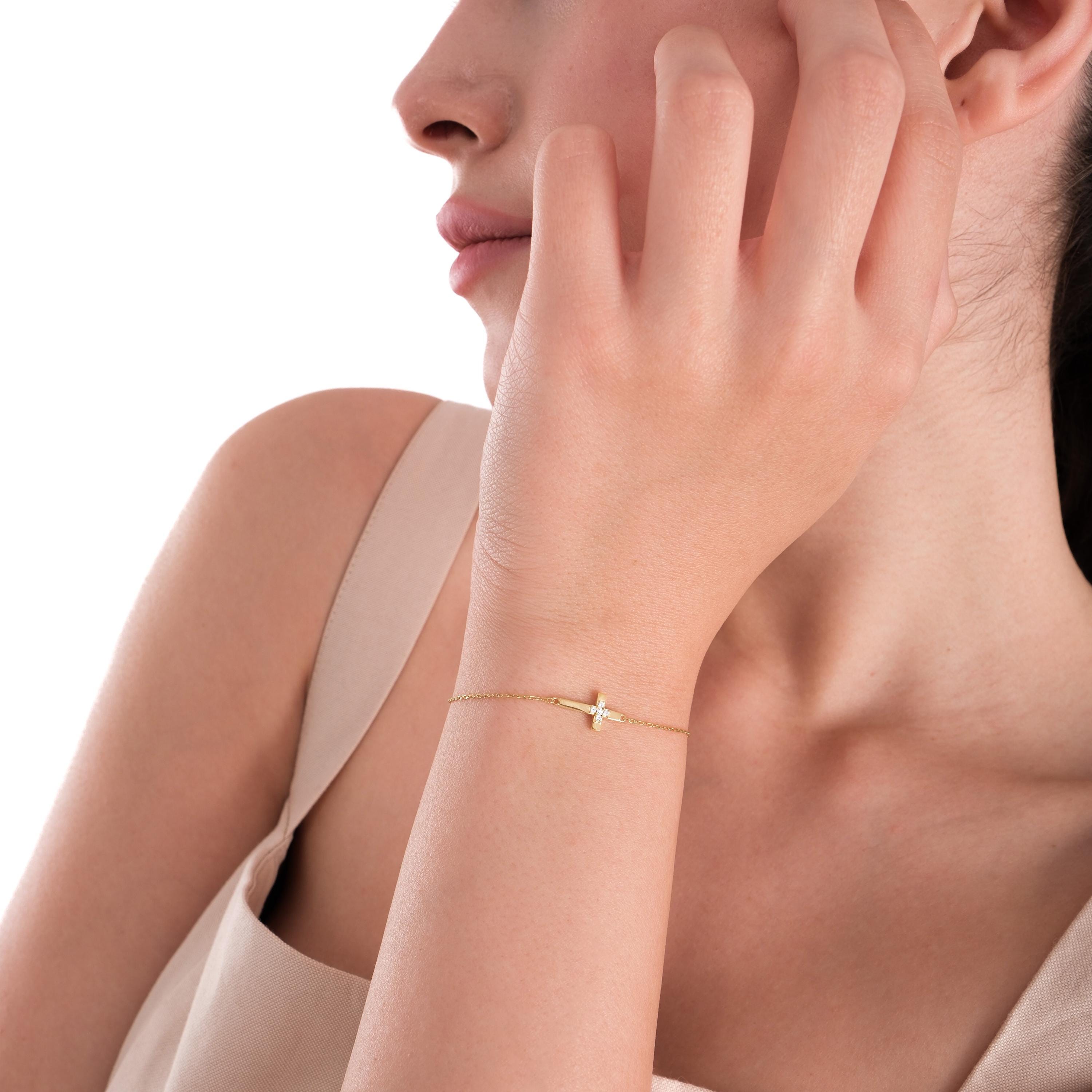 Side portrait of a woman showing off a 14K solid gold cross bracelet with diamonds, worn with a neutral-toned dress.